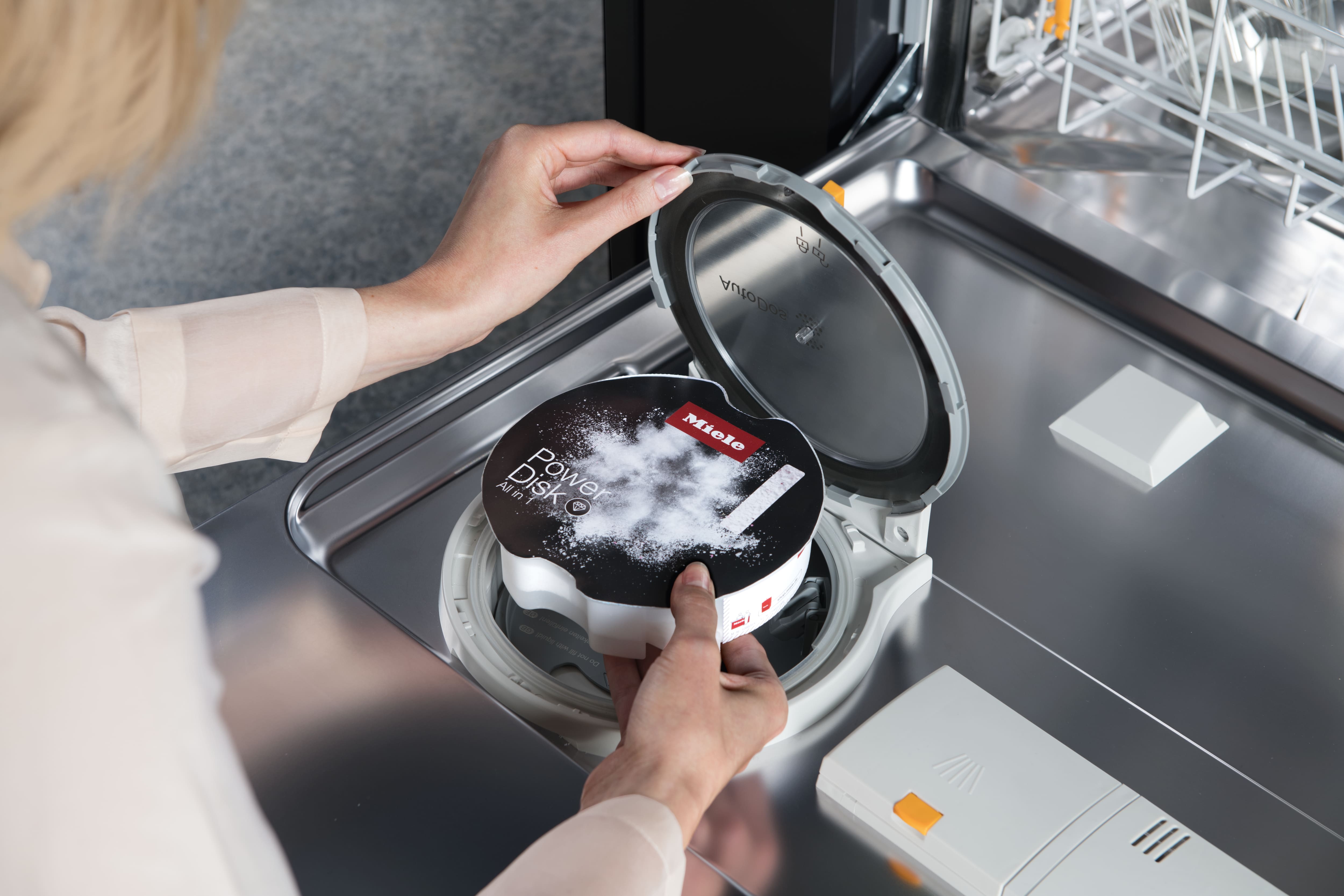 Person inserting a Miele PowerDisk detergent into a dishwasher’s AutoDos compartment for automatic dosing. Person inserting a Miele PowerDisk detergent into a dishwasher’s AutoDos compartment for automatic dosing.