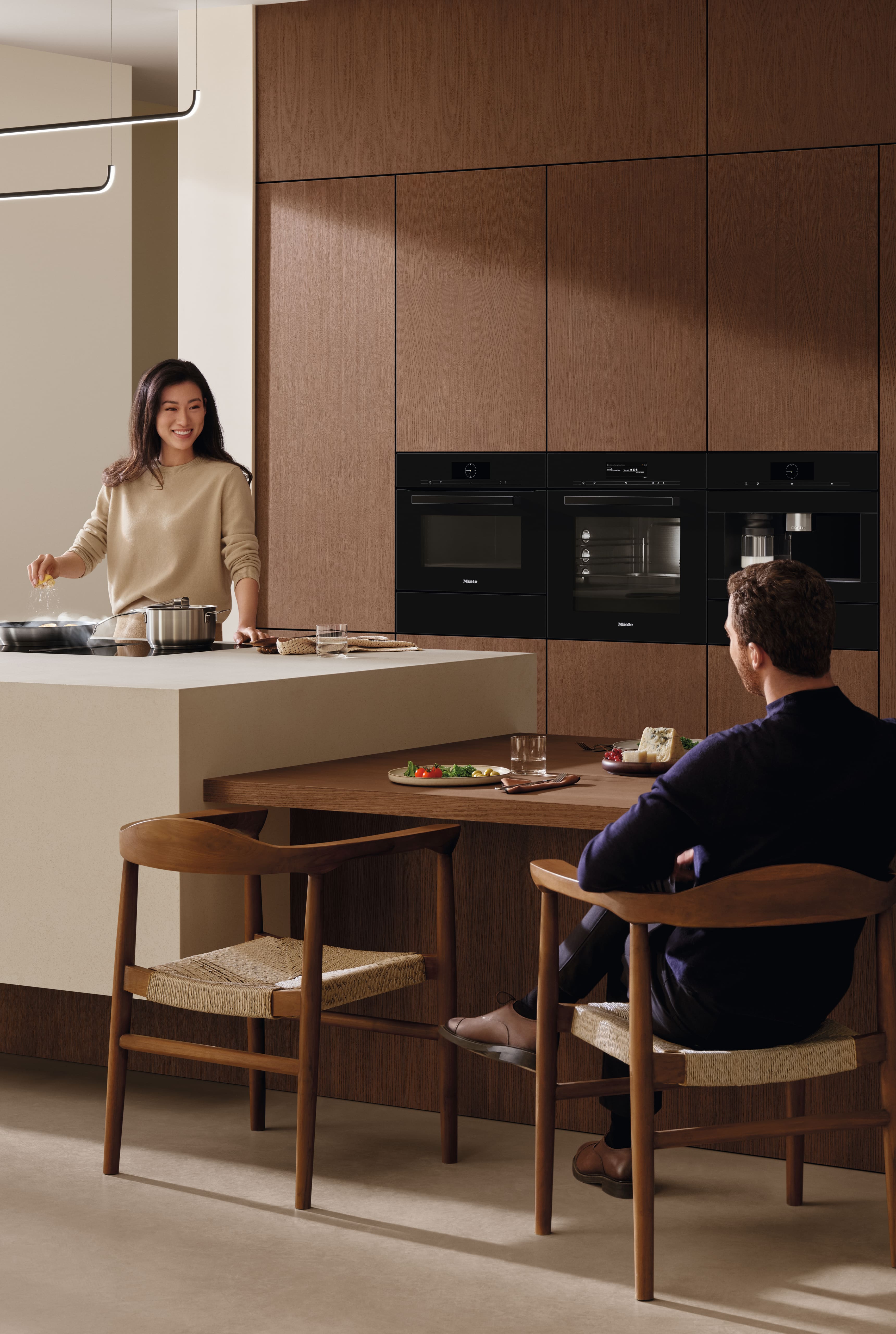 Cocina moderna con gabinetes de madera y electrodomésticos negros integrados; mujer cocinando en la isla, hombre sentado en el mostrador.
