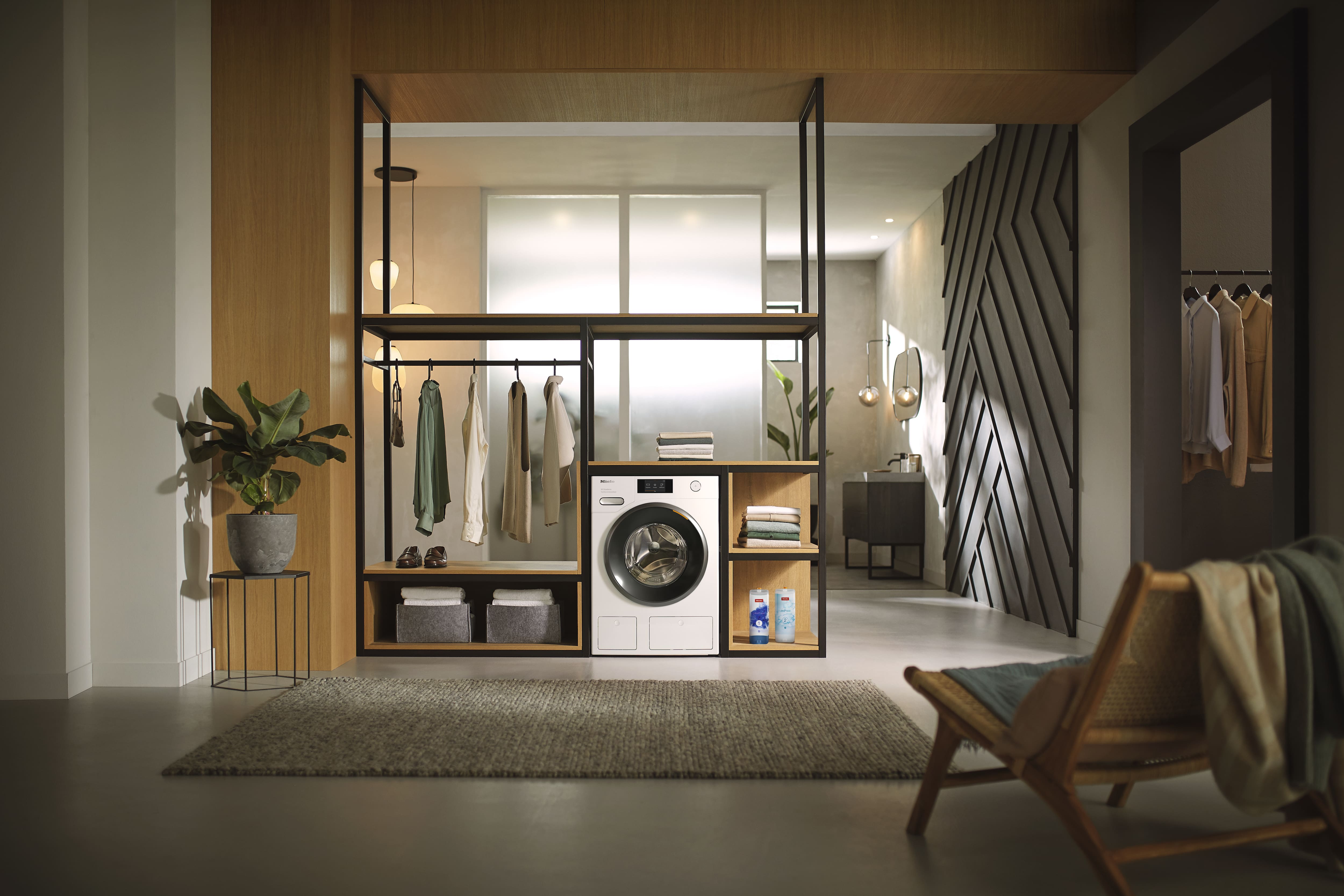 Modern open closet and laundry area with built-in front-loading washing machine, black-metal and wood shelving, hanging neutral clothing, folded towels, detergent bottles, a potted plant, and a rug and lounge chair in a softly lit contemporary interior. Modern open closet and laundry area with built-in front-loading washing machine, black-metal and wood shelving, hanging neutral clothing, folded towels, detergent bottles, a potted plant, and a rug and lounge chair in a softly lit contemporary interior.