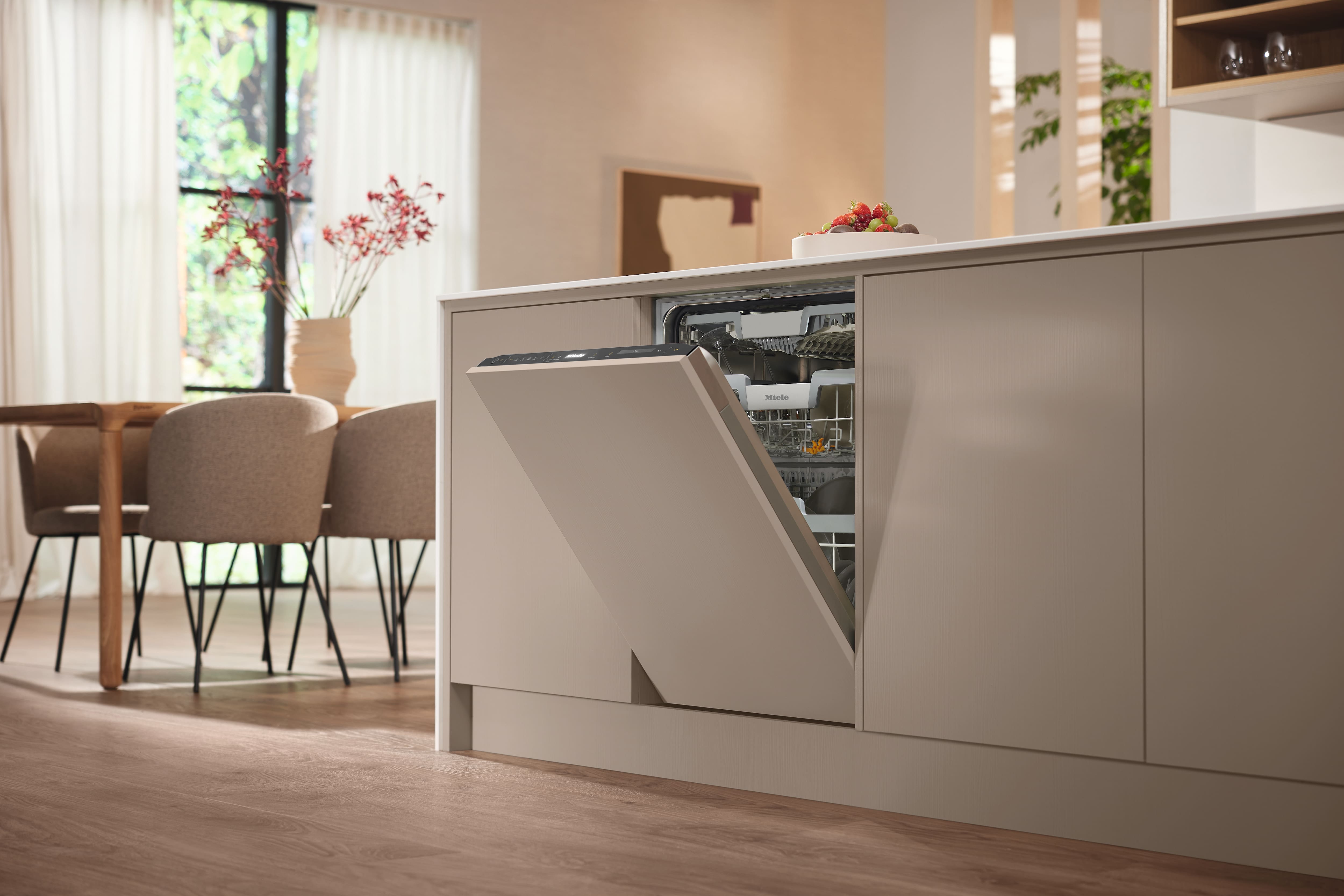 Cuisine moderne avec porte de lave-vaisselle intégrée entrouverte dans un mobilier beige ; coin repas et lumière du soleil en arrière-plan. Cuisine moderne avec porte de lave-vaisselle intégrée entrouverte dans un mobilier beige ; coin repas et lumière du soleil en arrière-plan.