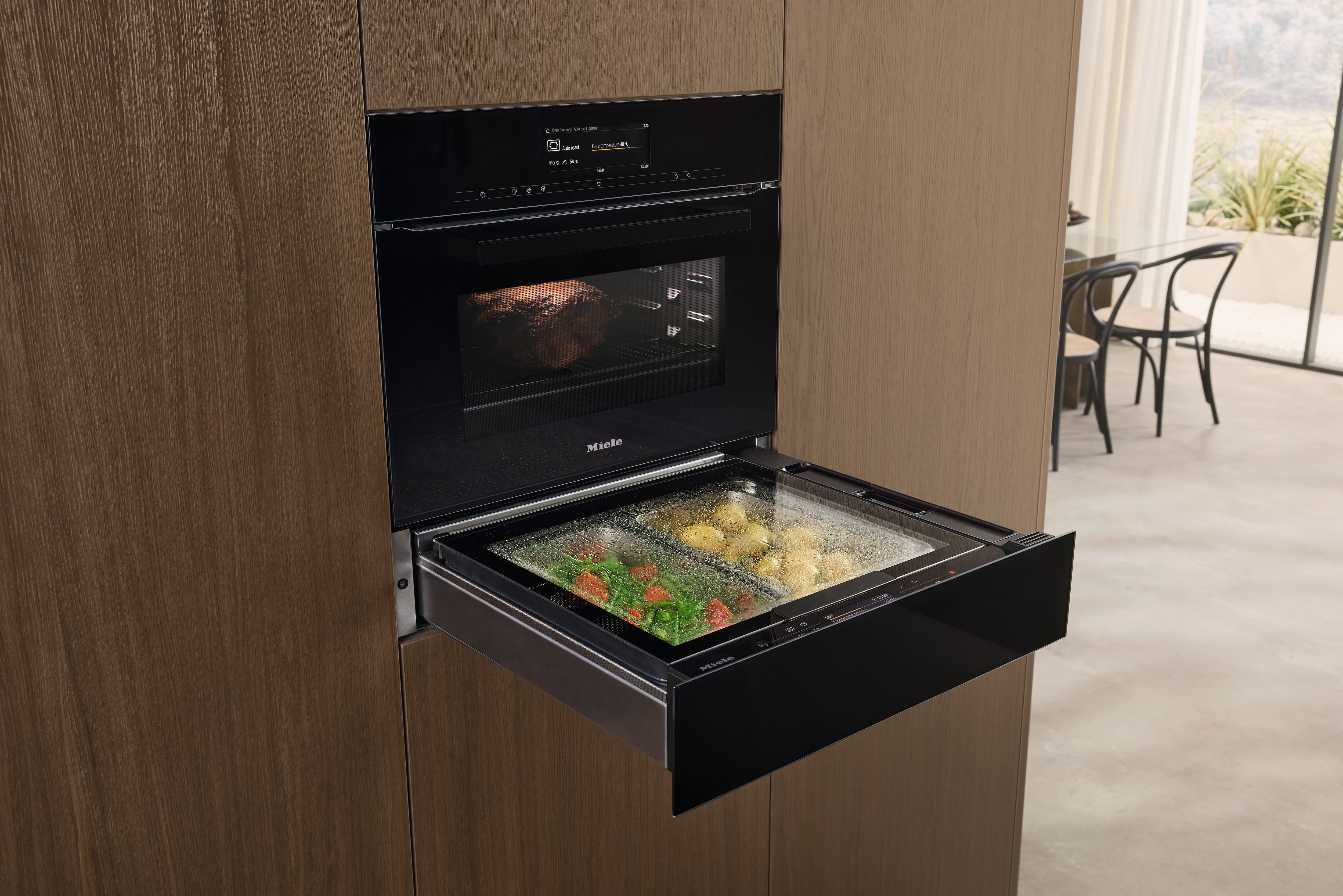 Moderne Küche mit eingebautem Miele-Ofen und offenem Dampfgarer, der Gemüse und Kartoffeln gart, während oben ein Braten gebacken wird. Moderne Küche mit eingebautem Miele-Ofen und offenem Dampfgarer, der Gemüse und Kartoffeln gart, während oben ein Braten gebacken wird.