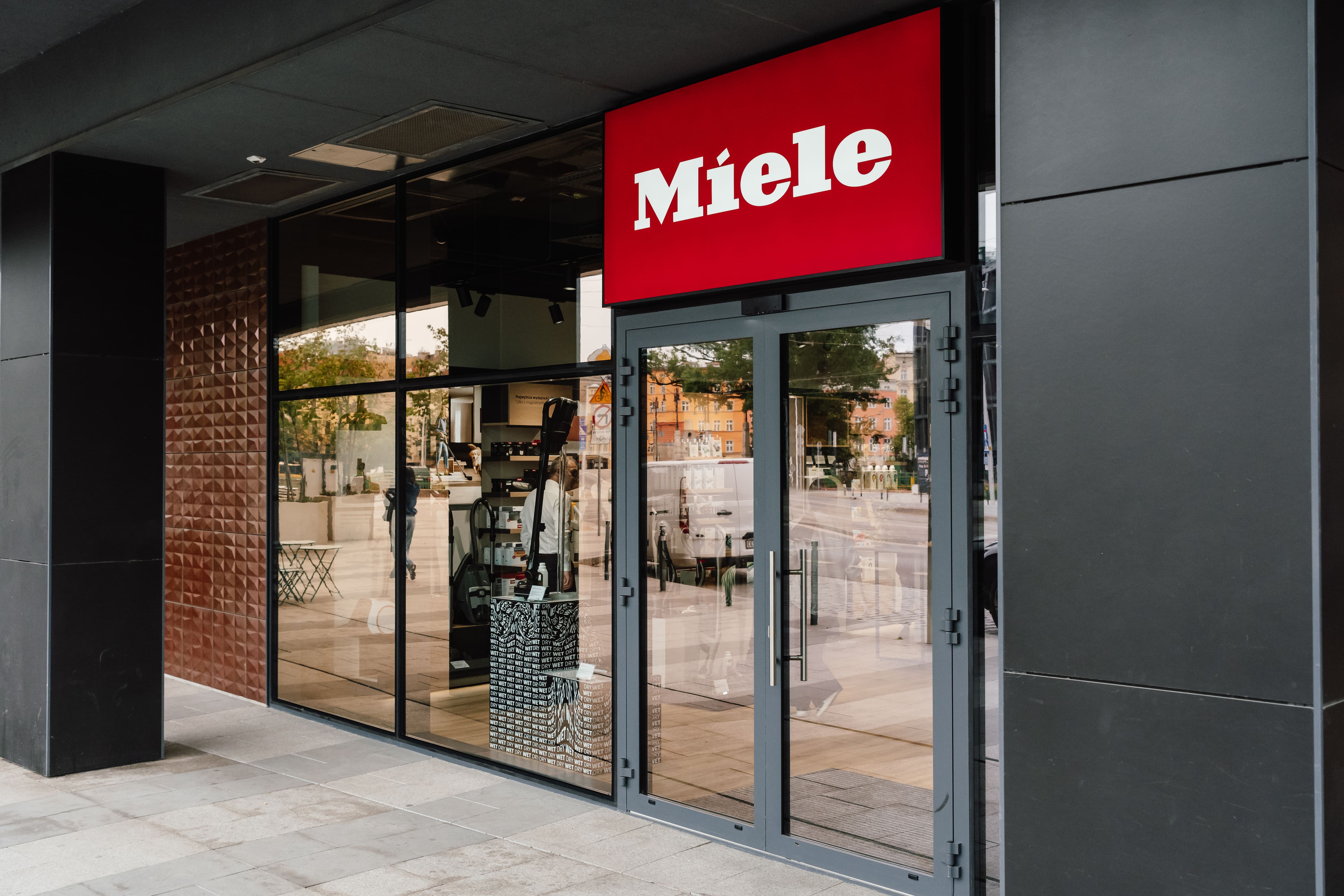 Street-level view of a Miele retail store entrance with red signage and glass facade showcasing appliances.