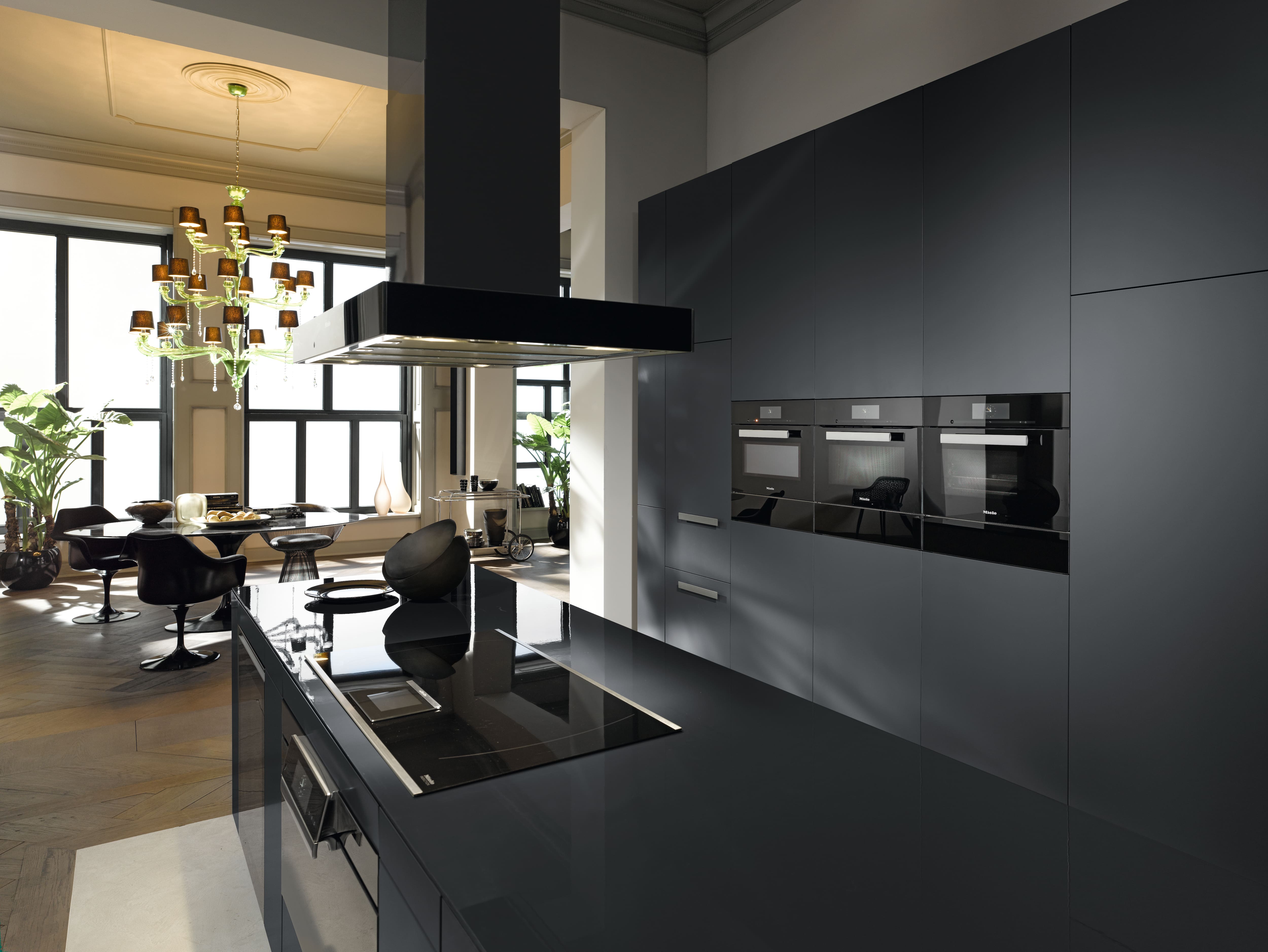 Sleek black kitchen with glossy island cooktop, wall ovens, dining nook and chandelier, showcasing luxury design. Sleek black kitchen with glossy island cooktop, wall ovens, dining nook and chandelier, showcasing luxury design.