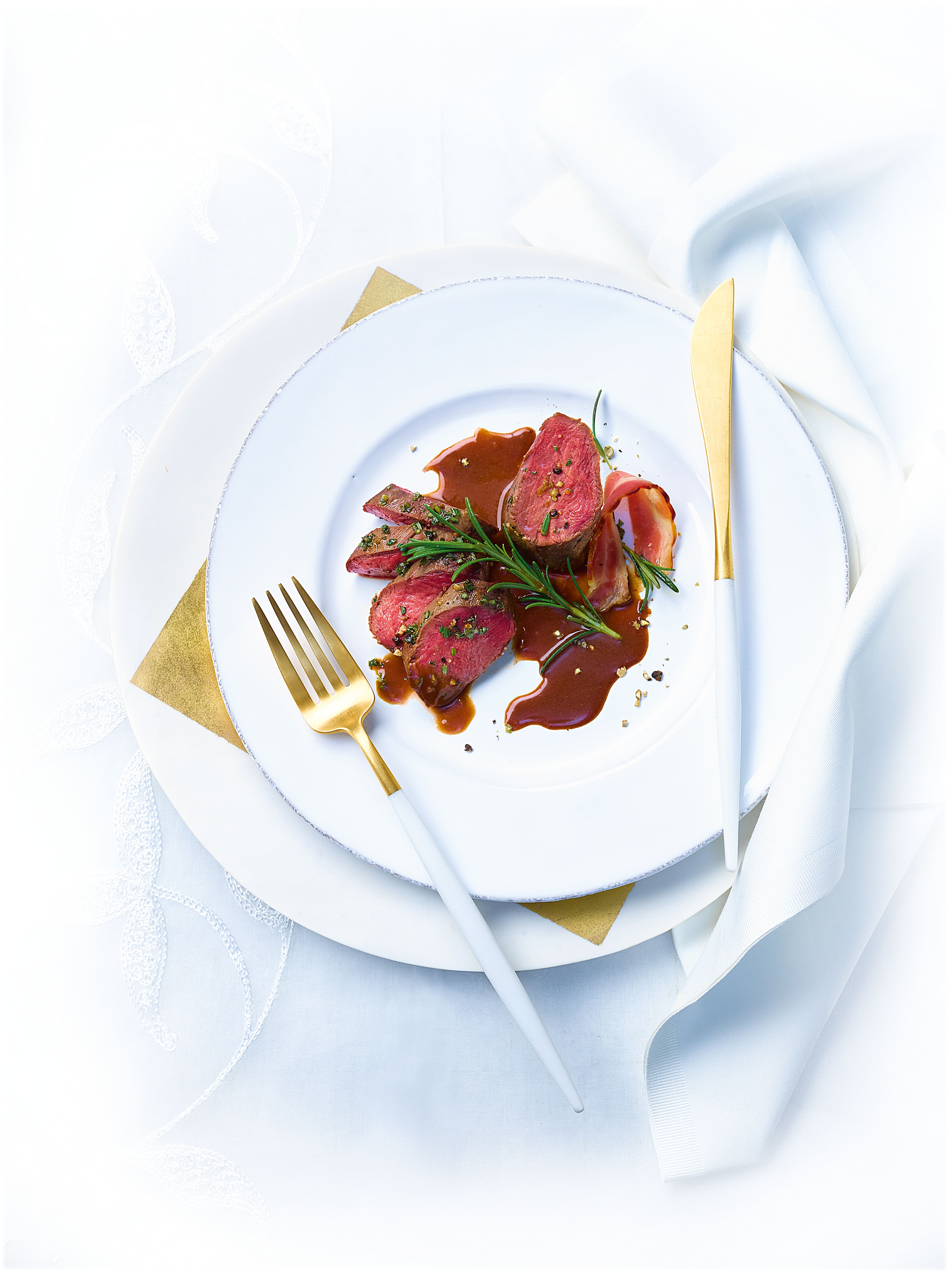 Fine-dining plate of medium-rare beef medallions with rosemary and glossy jus, prosciutto, and gold cutlery. Fine-dining plate of medium-rare beef medallions with rosemary and glossy jus, prosciutto, and gold cutlery.