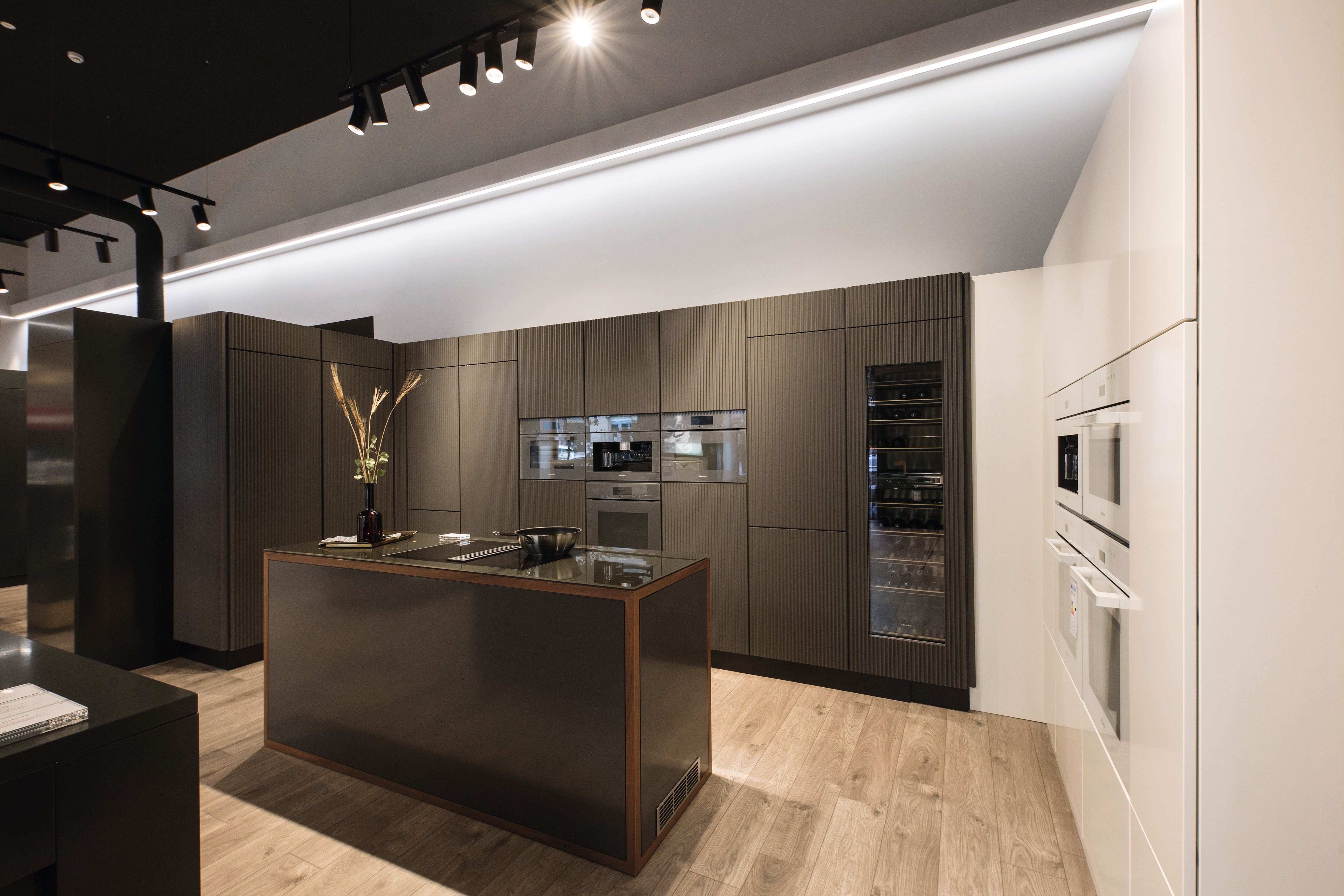 Cucina moderna con armadi scuri, piano cottura sull'isola, forni integrati, frigo per vini e illuminazione a binario. Cucina moderna con armadi scuri, piano cottura sull'isola, forni integrati, frigo per vini e illuminazione a binario.