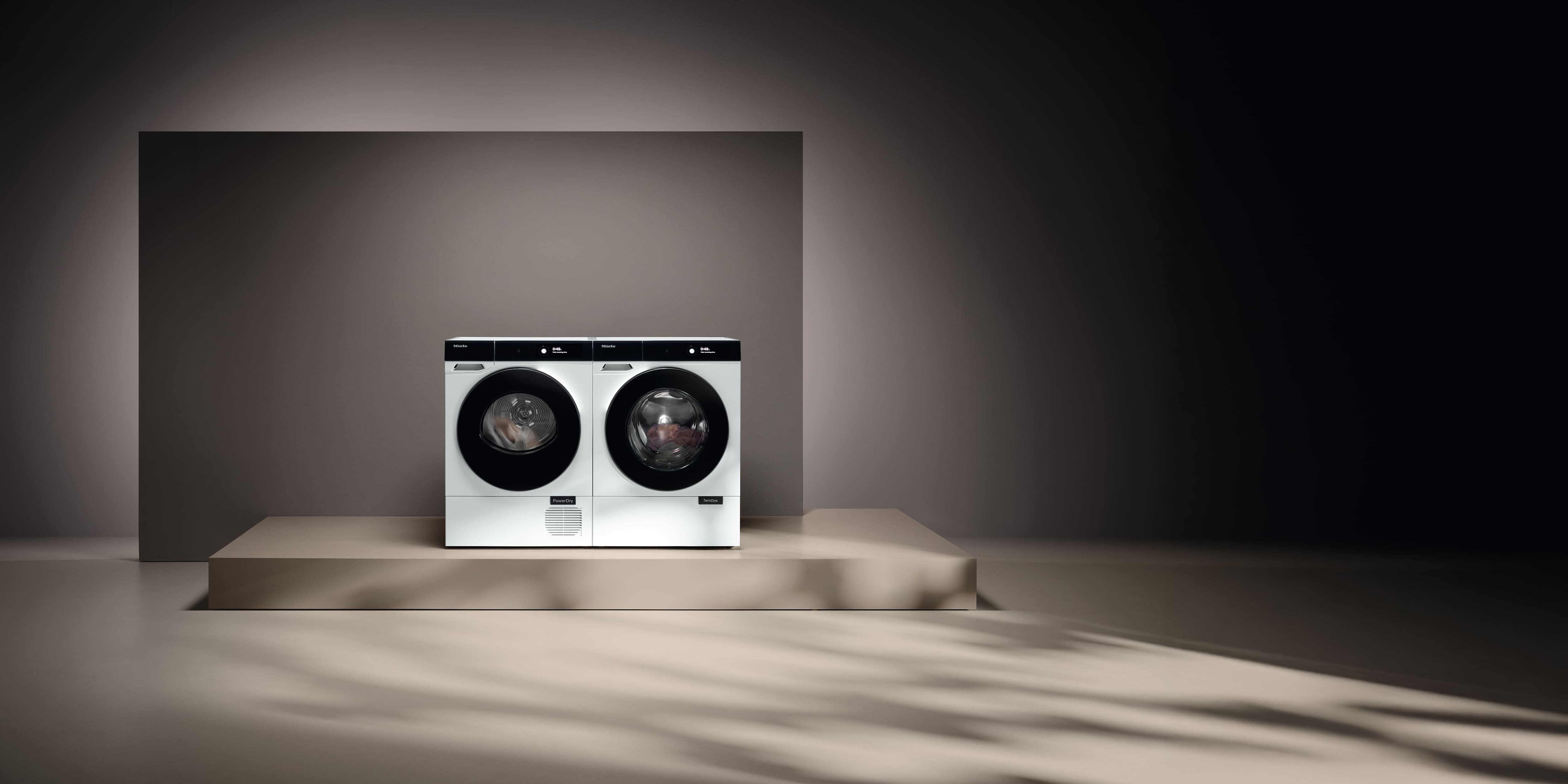 Minimalistisches Studio mit zwei weißen Miele-Frontlader-Waschmaschinen auf einem Podest, sanfte Schatten und neutraler Hintergrund.