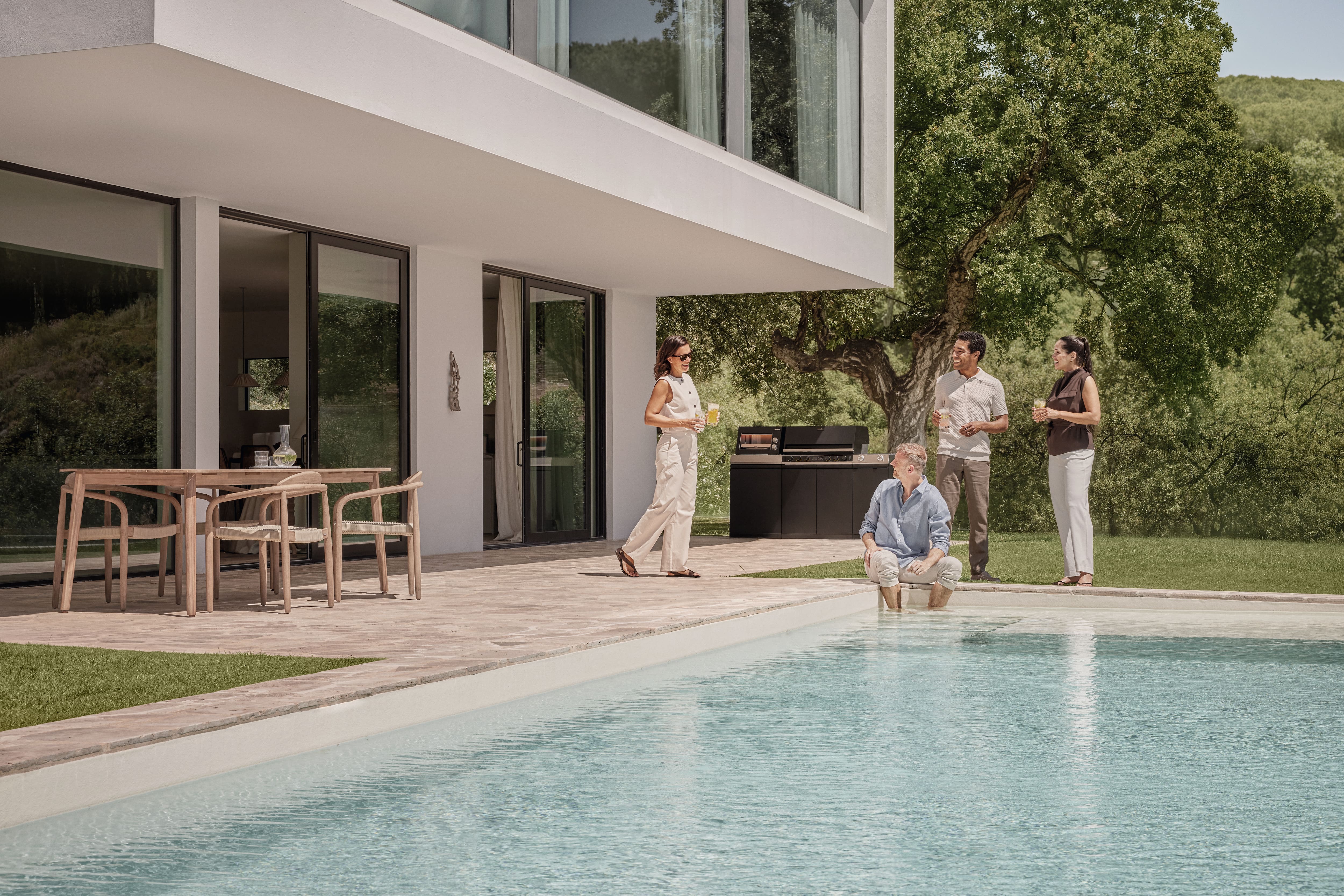 Moderne terrasse ved en pool, hvor fire voksne står og taler sammen nær en grill og et bord, omgivet af frodige træer på en solrig dag. Moderne terrasse ved en pool, hvor fire voksne står og taler sammen nær en grill og et bord, omgivet af frodige træer på en solrig dag.