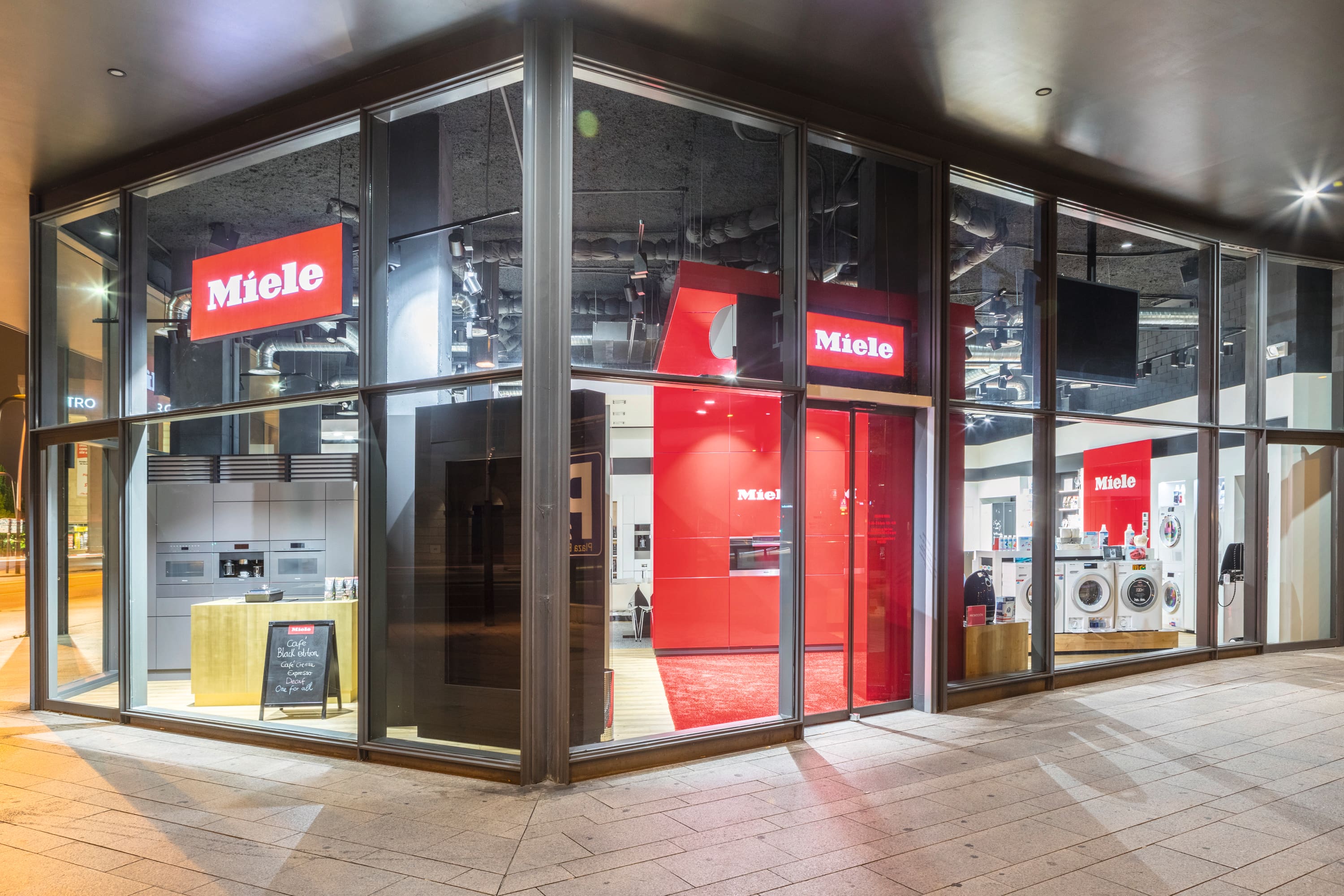 Showroom de electrodomésticos Miele con paredes de vidrio por la noche, mostrando lavadoras y cocinas integradas con audaz marca roja. Showroom de electrodomésticos Miele con paredes de vidrio por la noche, mostrando lavadoras y cocinas integradas con audaz marca roja.