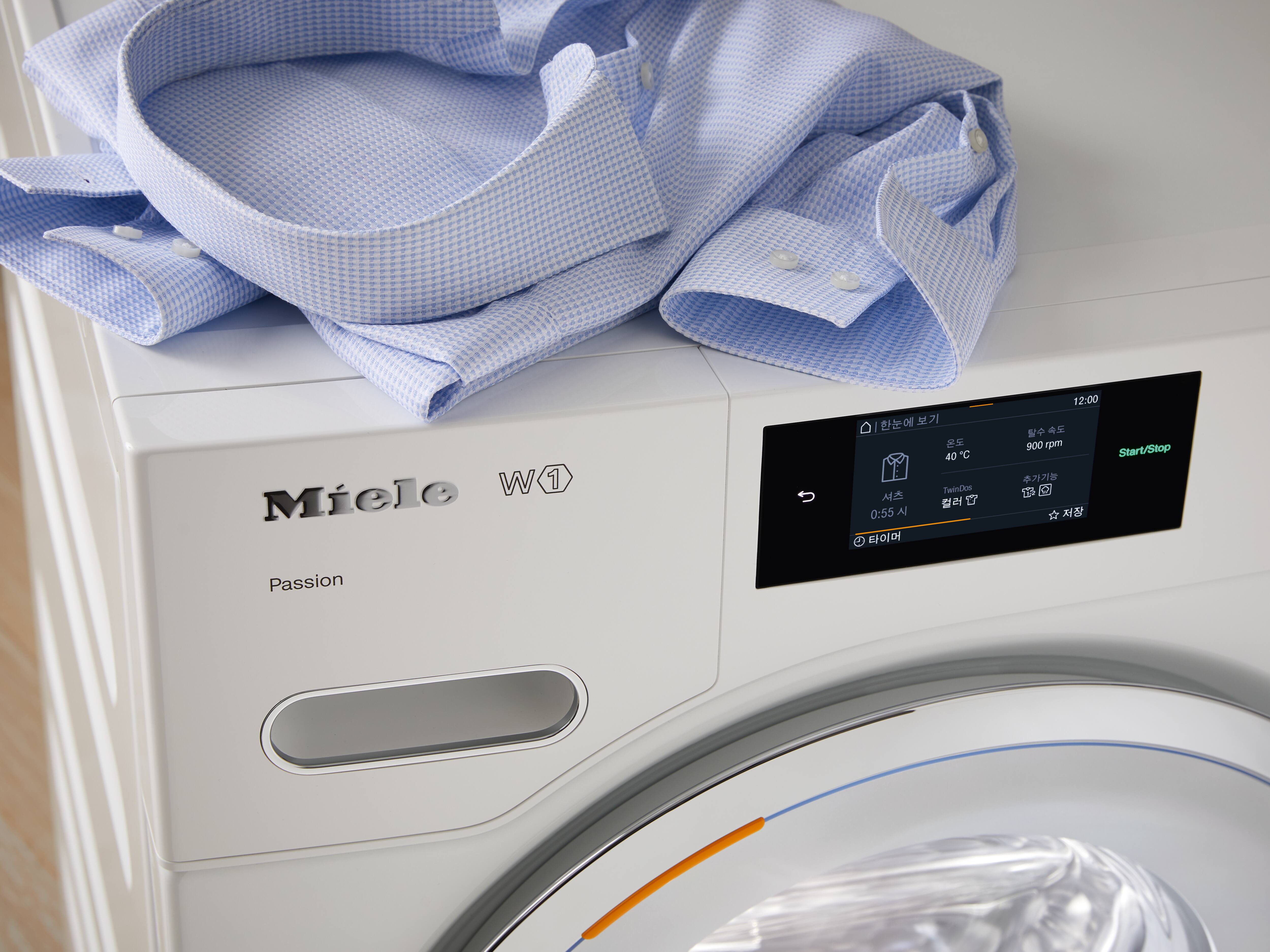 Close-up de uma lavadora Miele W1 branca com painel de controle digital e uma camisa cinza-escura colocada em cima; o texto na máquina menciona PowerWash, TwinDos e capacidade de 9 kg. Close-up de uma lavadora Miele W1 branca com painel de controle digital e uma camisa cinza-escura colocada em cima; o texto na máquina menciona PowerWash, TwinDos e capacidade de 9 kg.