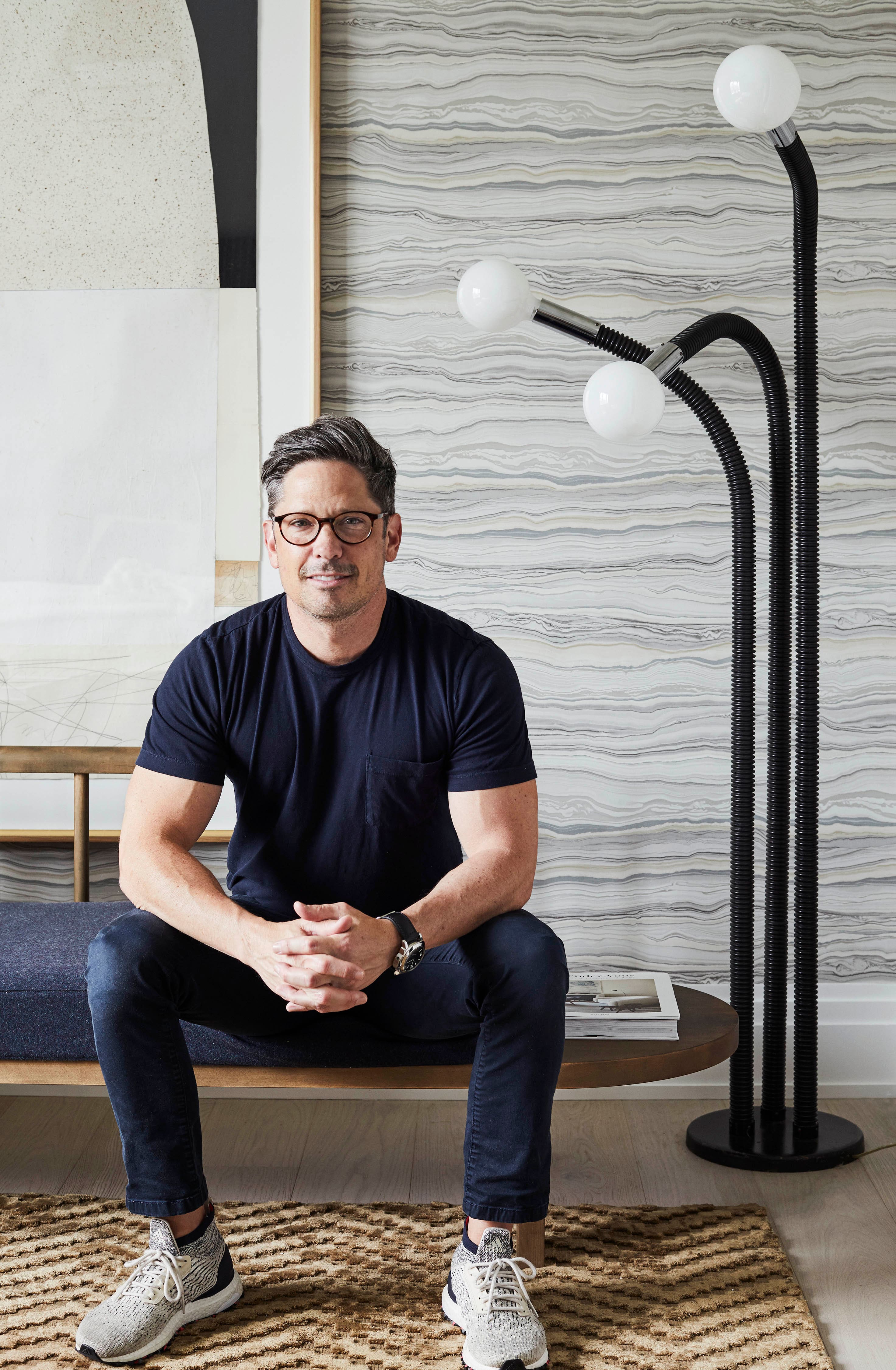 Man in casual black tee and glasses sits on bench in modern living room with abstract art and three-globe floor lamp.
