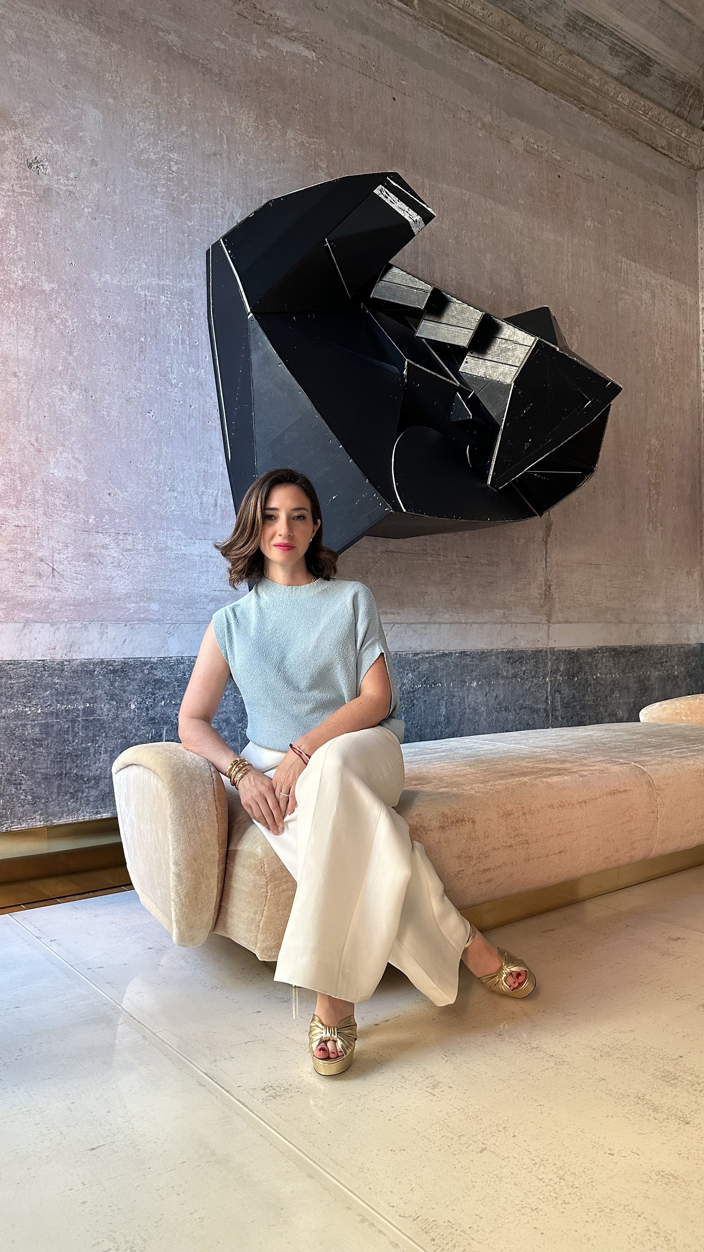 Person sits on a beige sofa in a modern room, wearing a light blue top and white trousers beneath a black geometric wall sculpture.