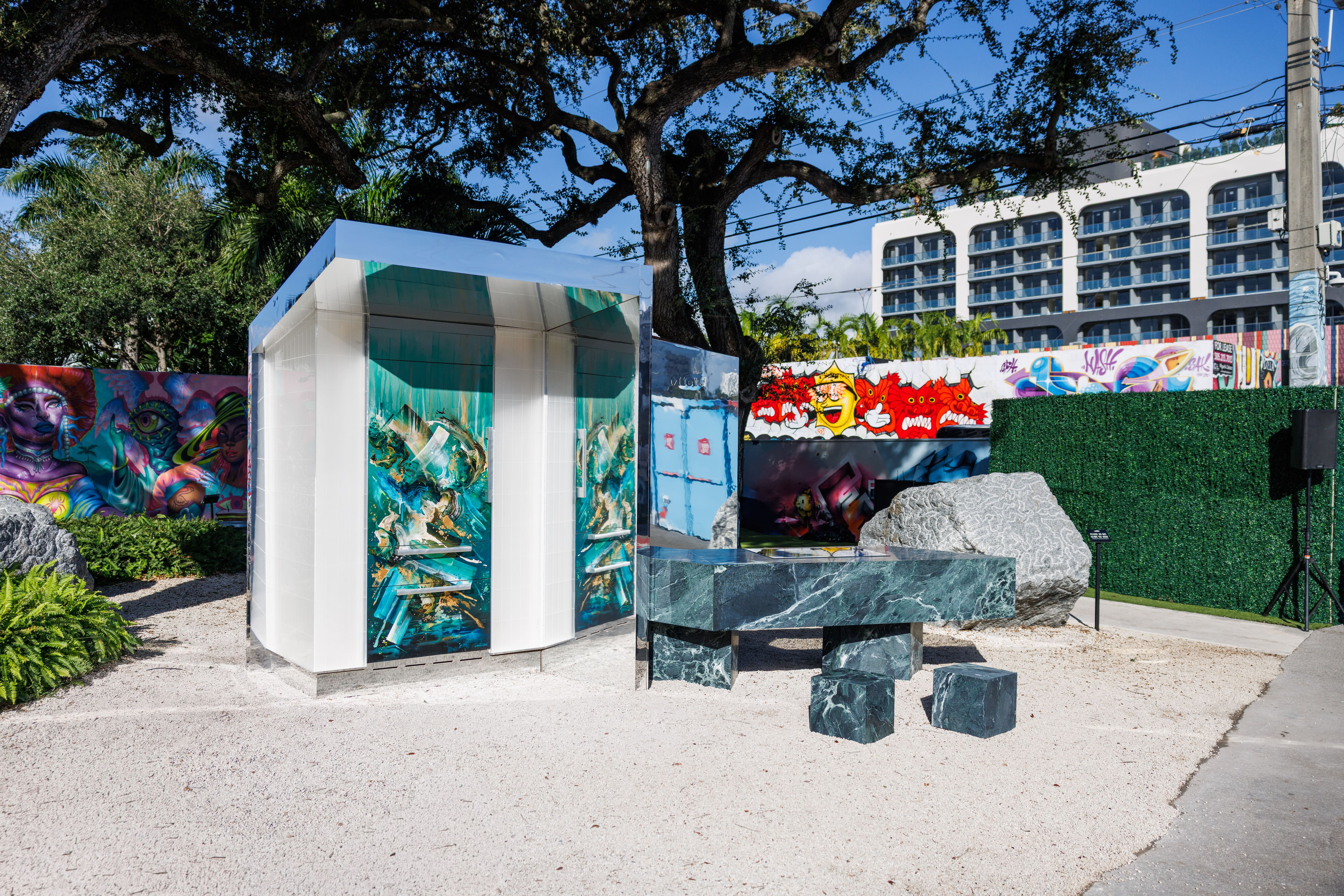 Outdoor art installation with a mirrored pavilion, green marble table, and colorful murals in a city courtyard. Outdoor art installation with a mirrored pavilion, green marble table, and colorful murals in a city courtyard.