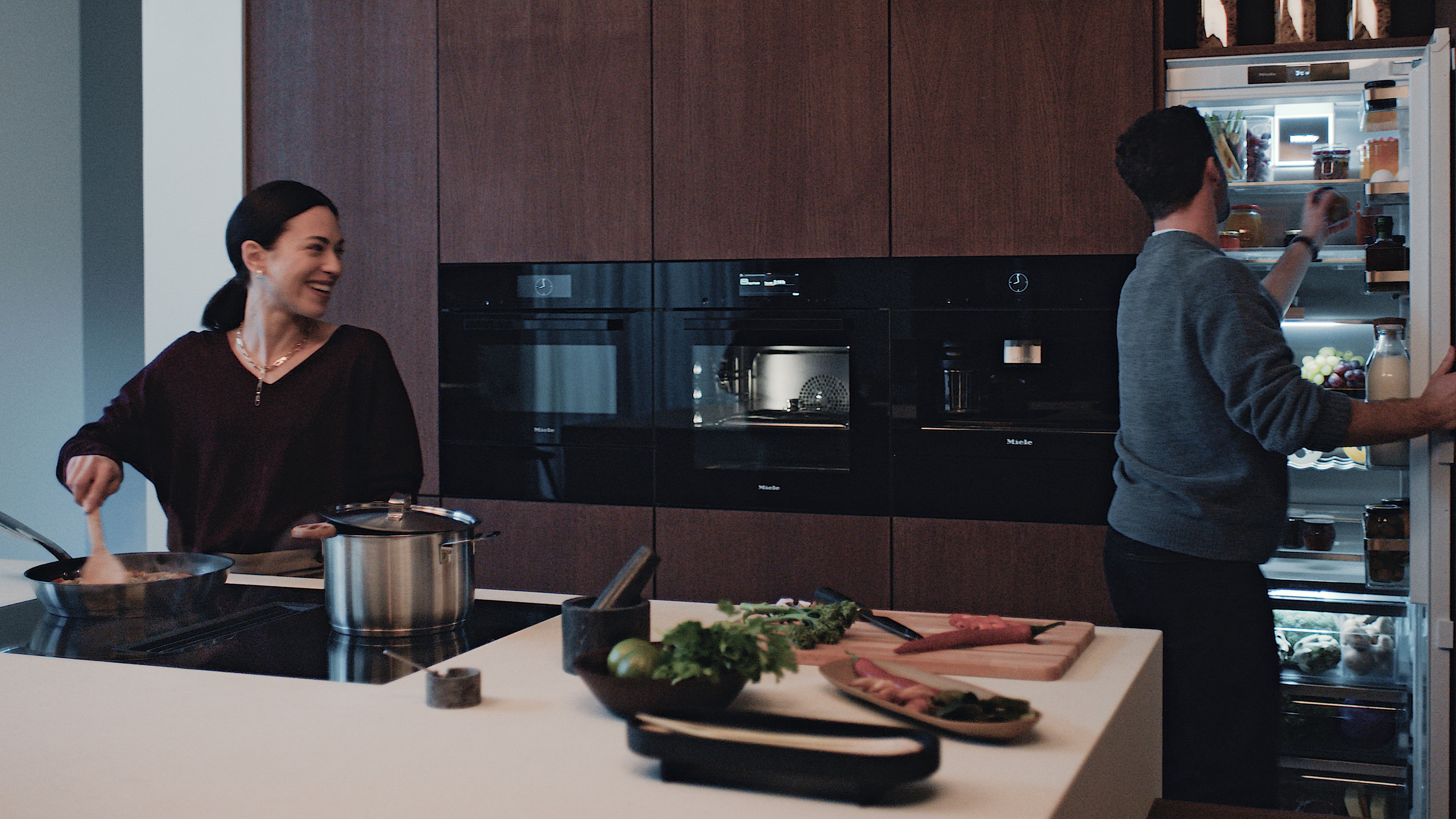 Kvinna lagar mat i ett kök med Mieleprodukter. En man plockar mat ut kylskåpet. Kvinna lagar mat i ett kök med Mieleprodukter. En man plockar mat ut kylskåpet.