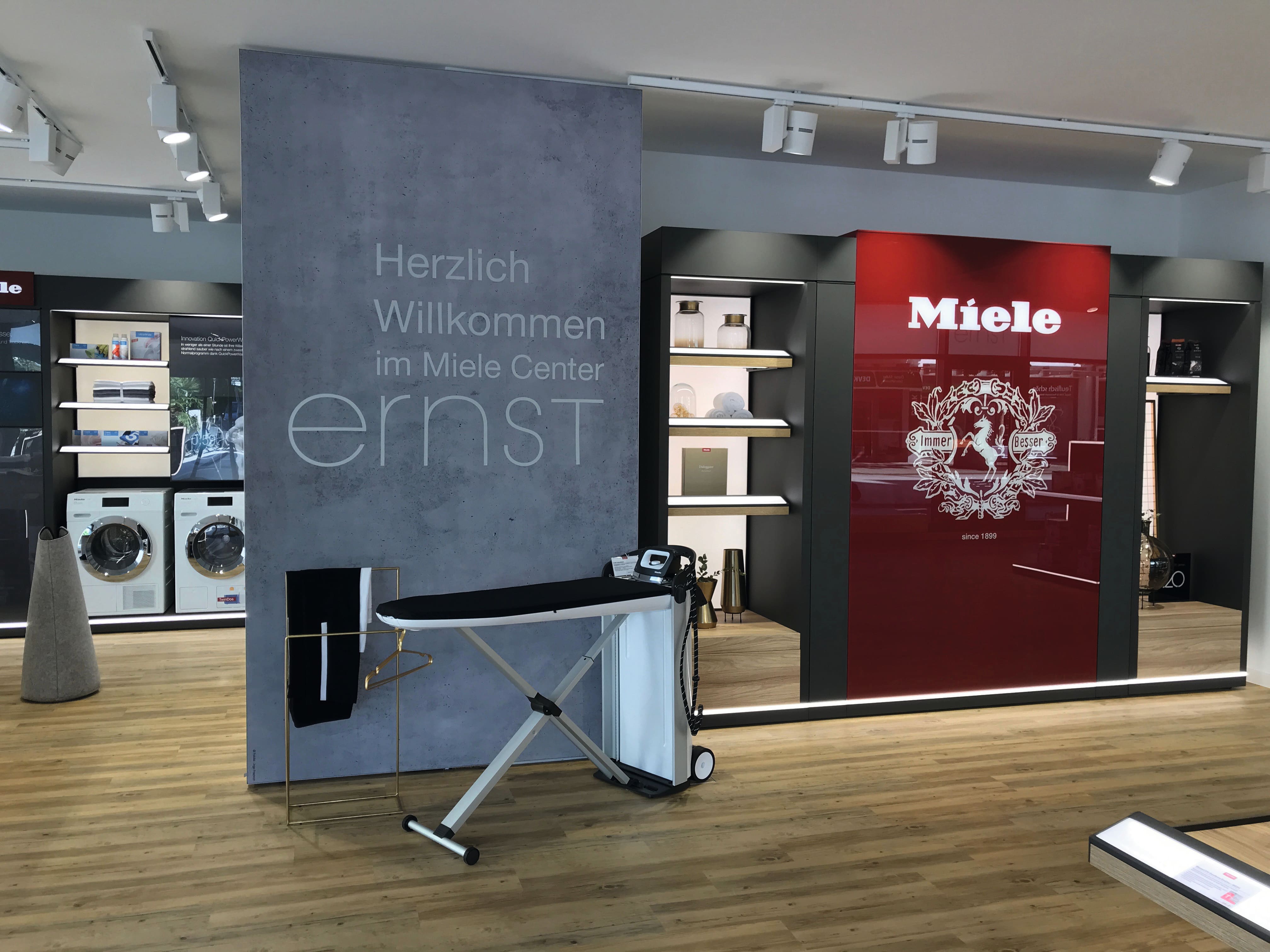 Moderner Miele-Showroom mit Waschmaschinen, Regalen, rotem Markenpanel und Bügelstation; Deutsches Willkommensschild.