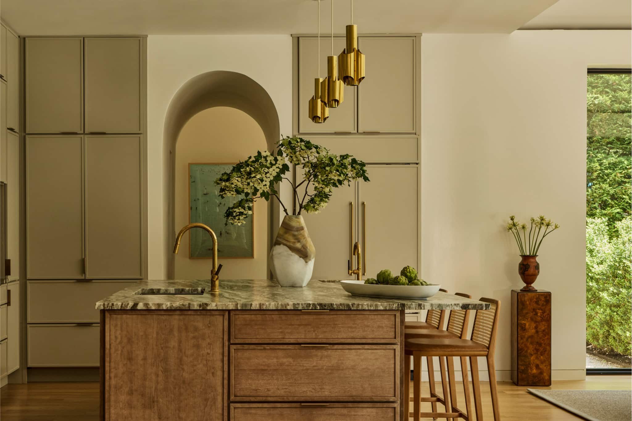 Warm modern kitchen with marble island, brass faucet and pendants, integrated cabinetry, and wooden stools.