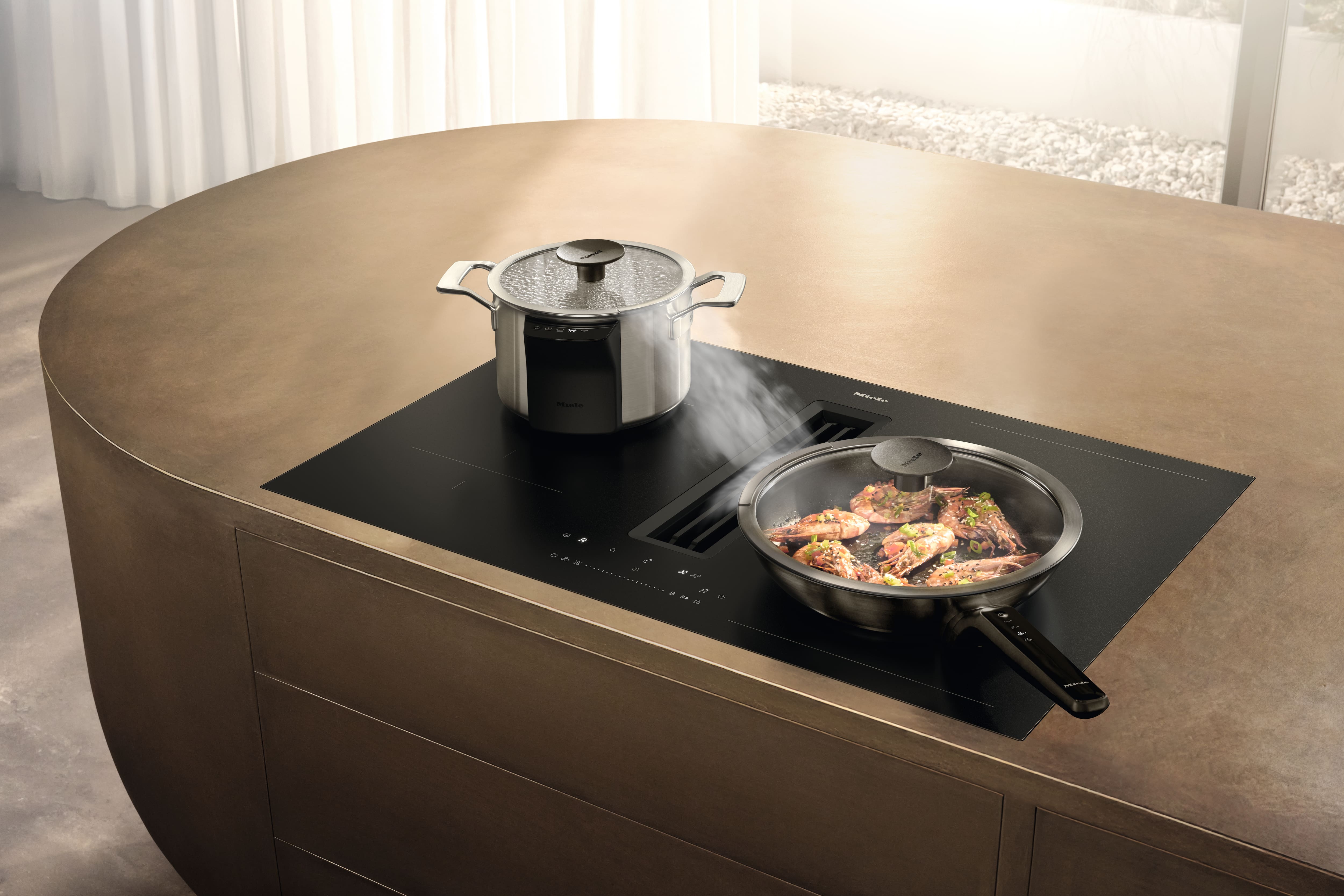 Plaque à induction élégante avec système de ventilation par aspiration vers le bas intégré, installée sur un îlot, permettant de cuisiner des plats et des casseroles à la vapeur.