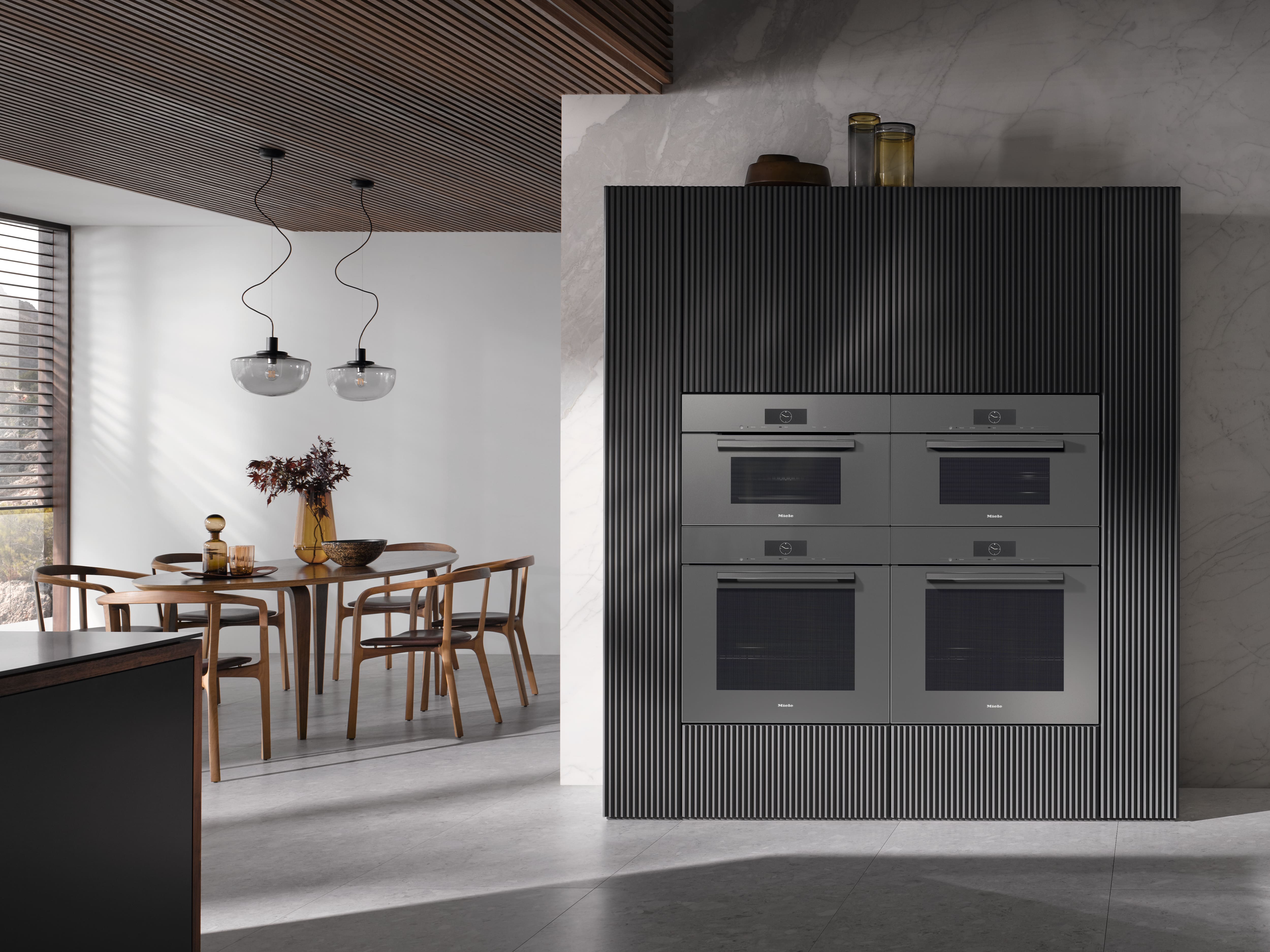 Modern kitchen with four built-in stainless ovens in a ribbed wall, wood dining table, and pendant lights. Modern kitchen with four built-in stainless ovens in a ribbed wall, wood dining table, and pendant lights.