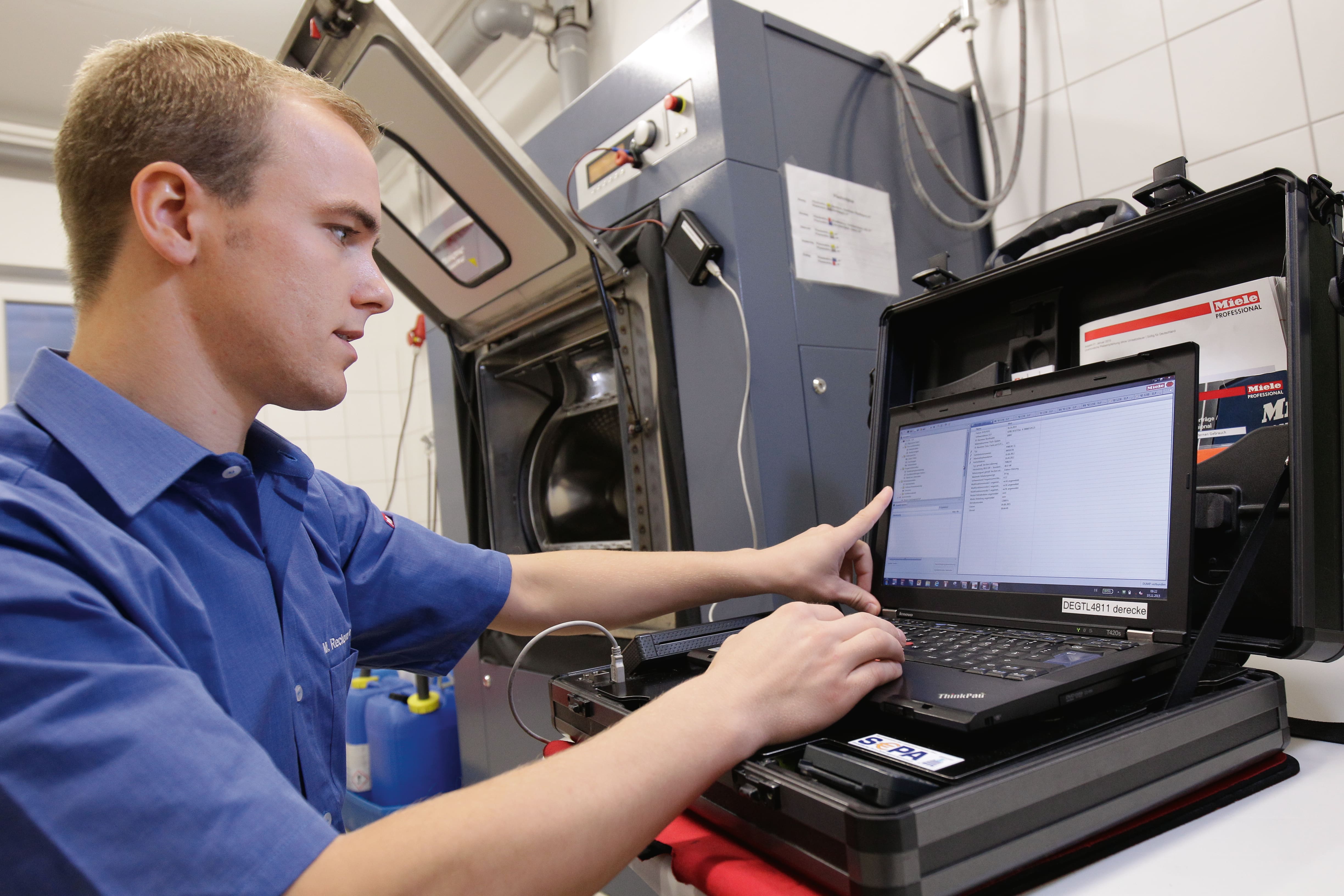 Technician using a laptop to diagnose and program an industrial washing machine during maintenance in a facility. Technician using a laptop to diagnose and program an industrial washing machine during maintenance in a facility.