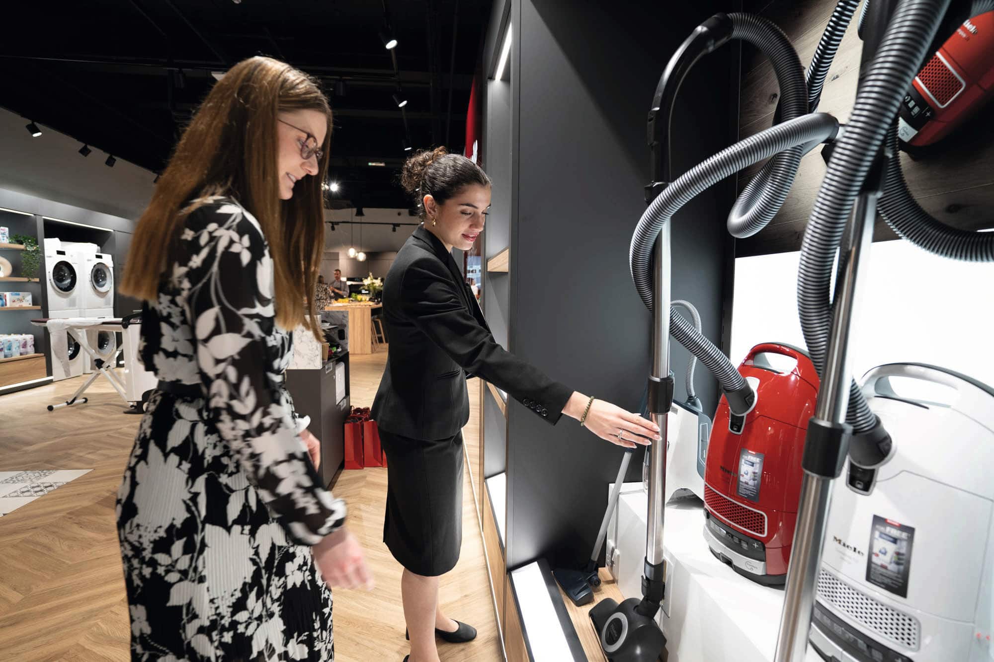 Two people in an appliance store examine canister vacuum cleaners on a display wall. A salesperson in a black suit gestures toward a vacuum while a customer in a patterned dress watches. Red and white vacuums are showcased; the showroom has wood floors and other appliances in the background.