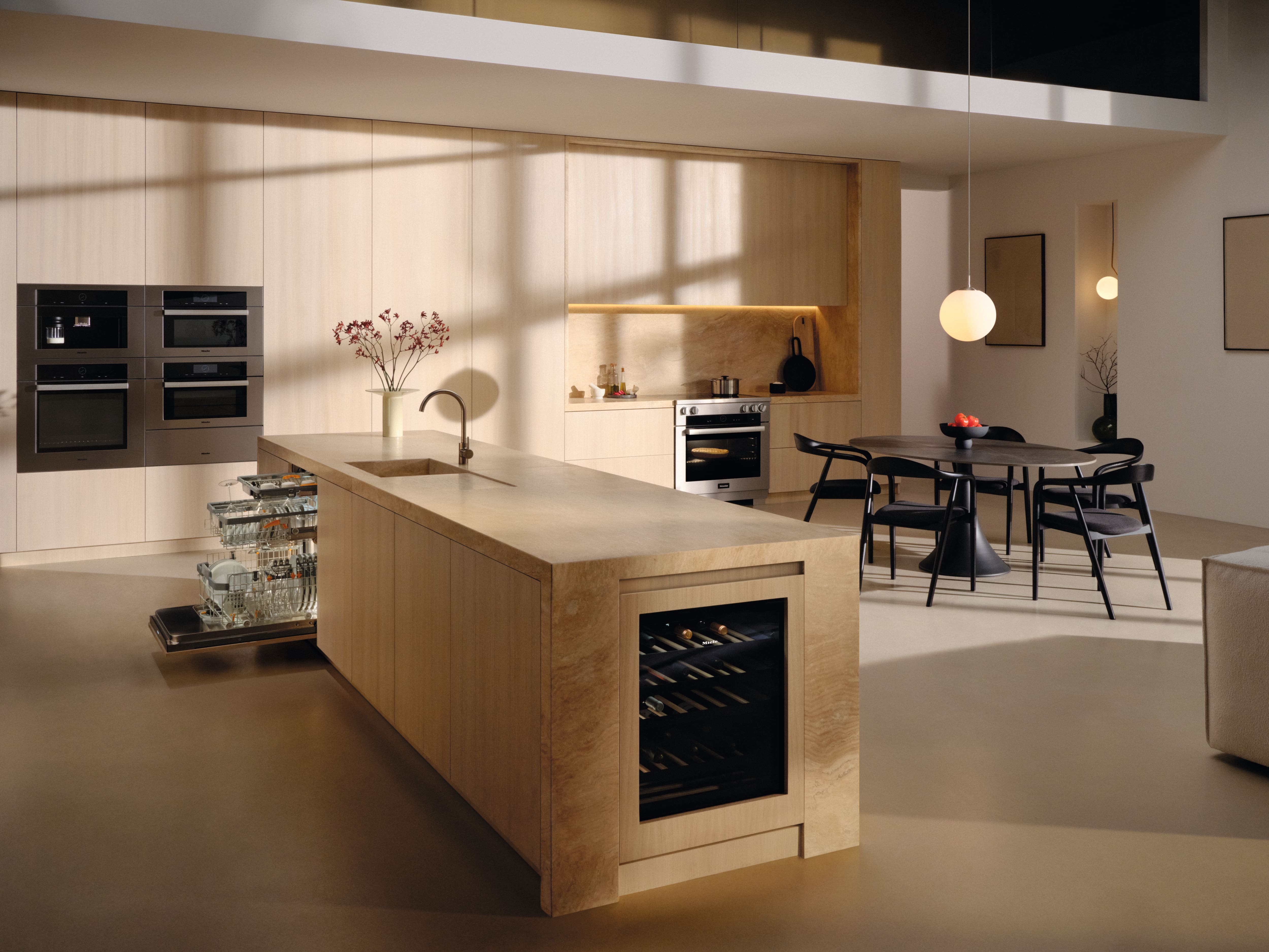 Minimalist kitchen with light wood cabinetry, island sink, open dishwasher, and dining area in warm light. Minimalist kitchen with light wood cabinetry, island sink, open dishwasher, and dining area in warm light.