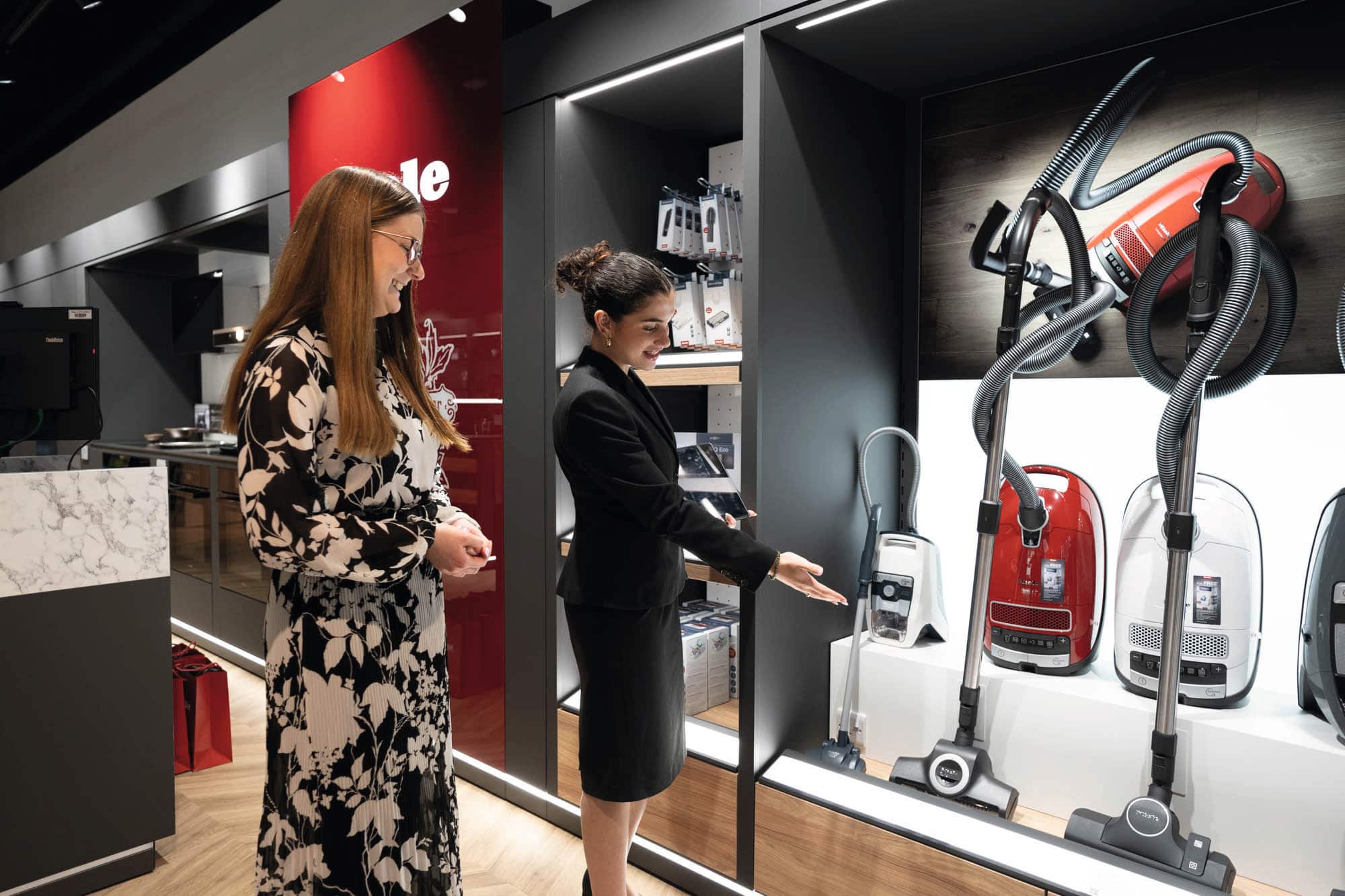 Two people in an appliance showroom looking at a display of canister vacuum cleaners; one person in a black suit gestures toward red and white vacuums with long hoses, with store shelves and product boxes in the background; faces blurred. Two people in an appliance showroom looking at a display of canister vacuum cleaners; one person in a black suit gestures toward red and white vacuums with long hoses, with store shelves and product boxes in the background; faces blurred.