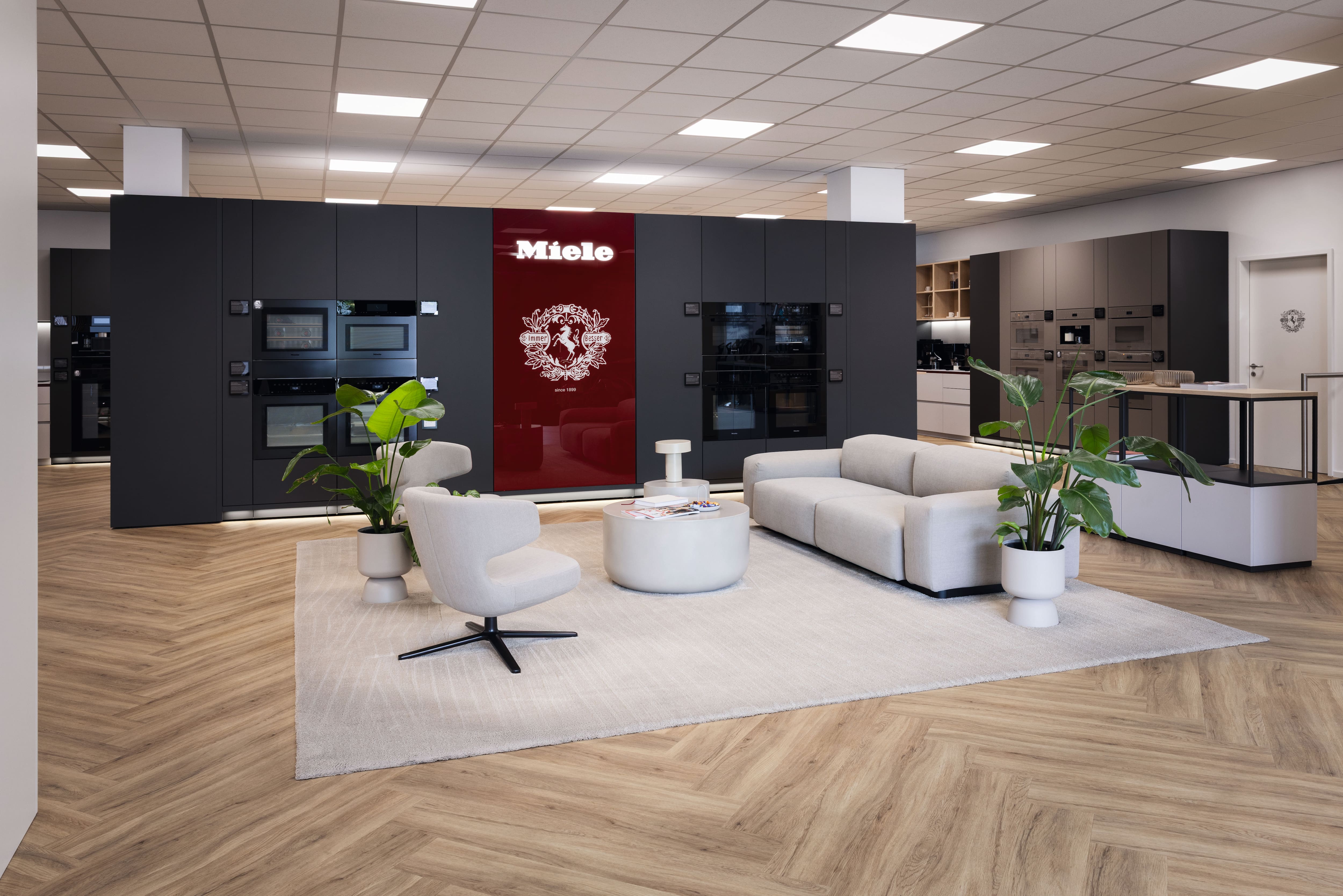 Moderner Showroom mit Miele Einbauküchengeräten in dunklen Schränken mit rotem Miele-Panel; helles Holzfischgrätparkett, neutrale Loungemöbel, Couchtisch und Topfpflanzen unter hellen Deckenleuchten.