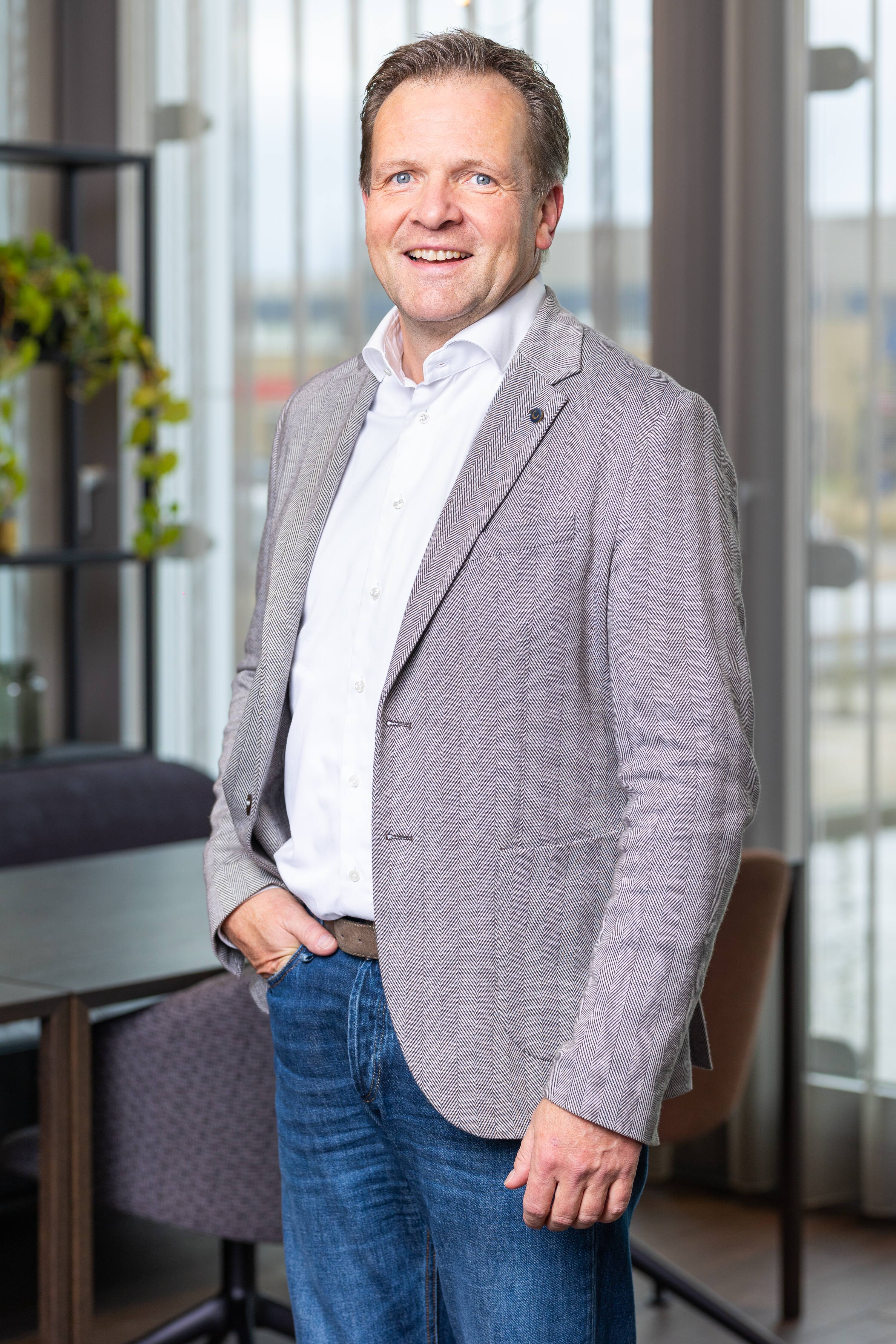 Business-casual portrait: man in gray herringbone blazer, white shirt, and jeans in a modern office setting.