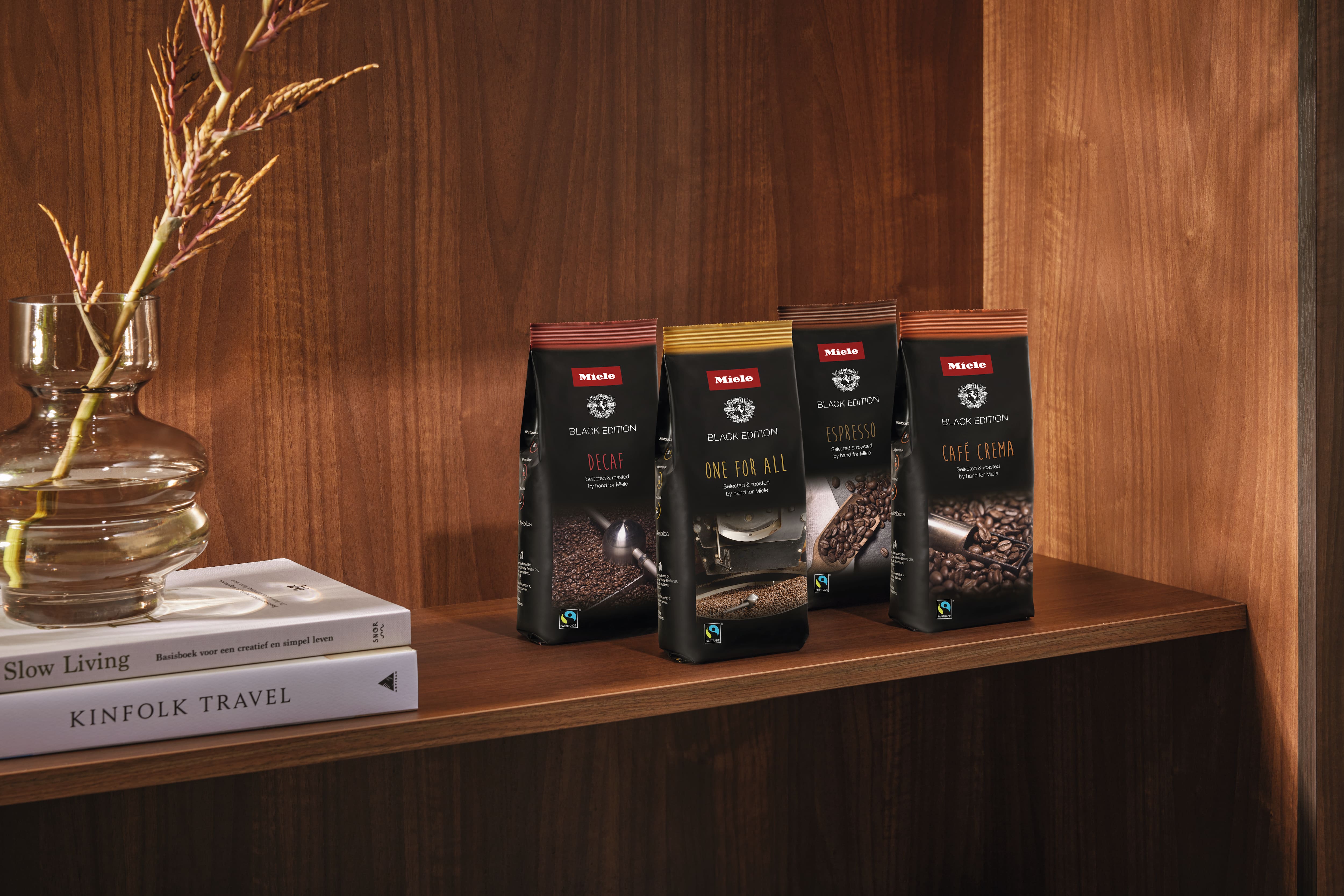Four Miele Black Edition coffee bags—Decaf, One for All, Espresso, Café Crema—on a wooden shelf with books and vase. Four Miele Black Edition coffee bags—Decaf, One for All, Espresso, Café Crema—on a wooden shelf with books and vase.