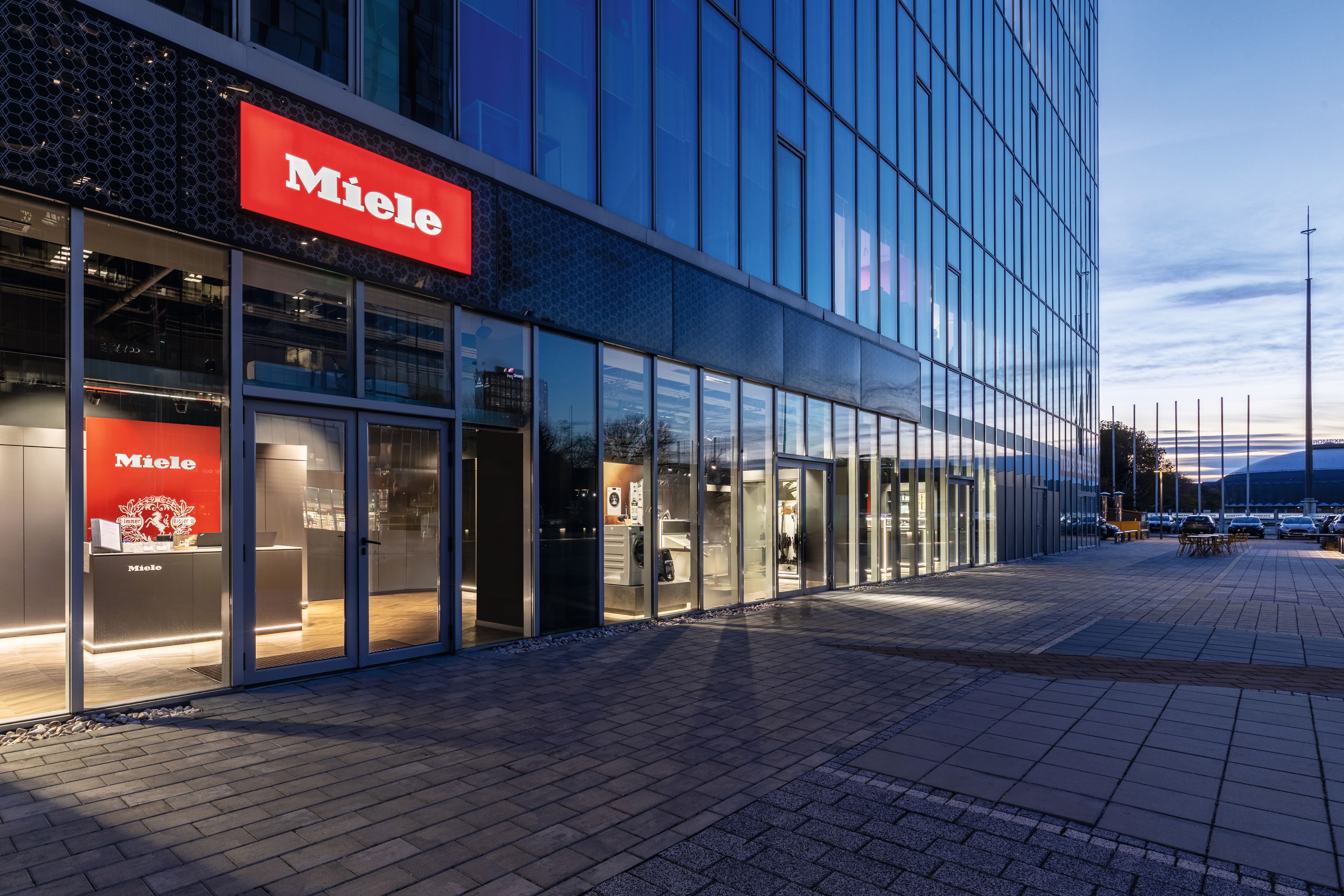 Clădire modernă din sticlă cu showroom Miele iluminat la amurg, prezentând aparate prin ferestre mari. Clădire modernă din sticlă cu showroom Miele iluminat la amurg, prezentând aparate prin ferestre mari.