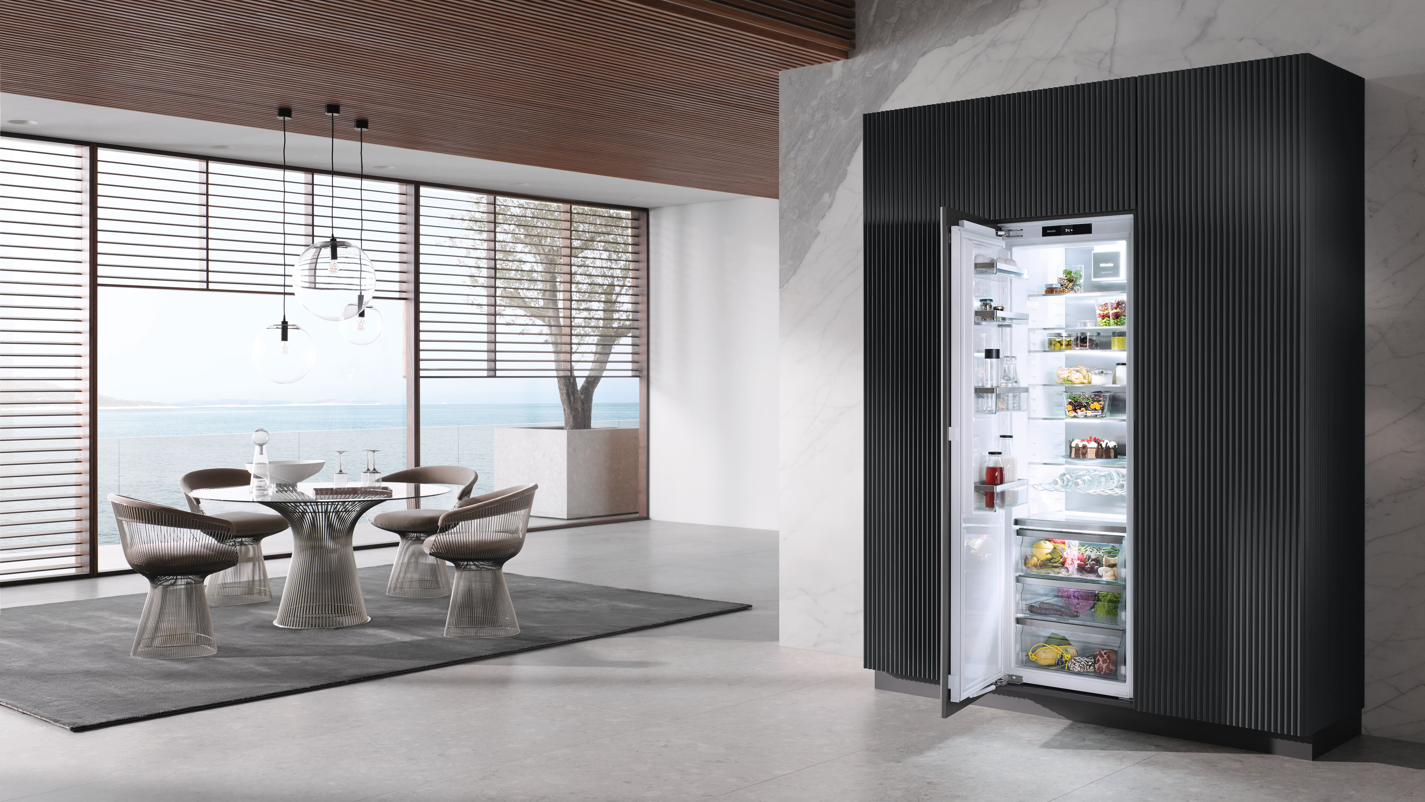Modern coastal dining area with large windows and wood‑slat ceiling, round wire-frame table and chairs under globe pendants, and an open built-in refrigerator set into a dark paneled wall beside a marble column.