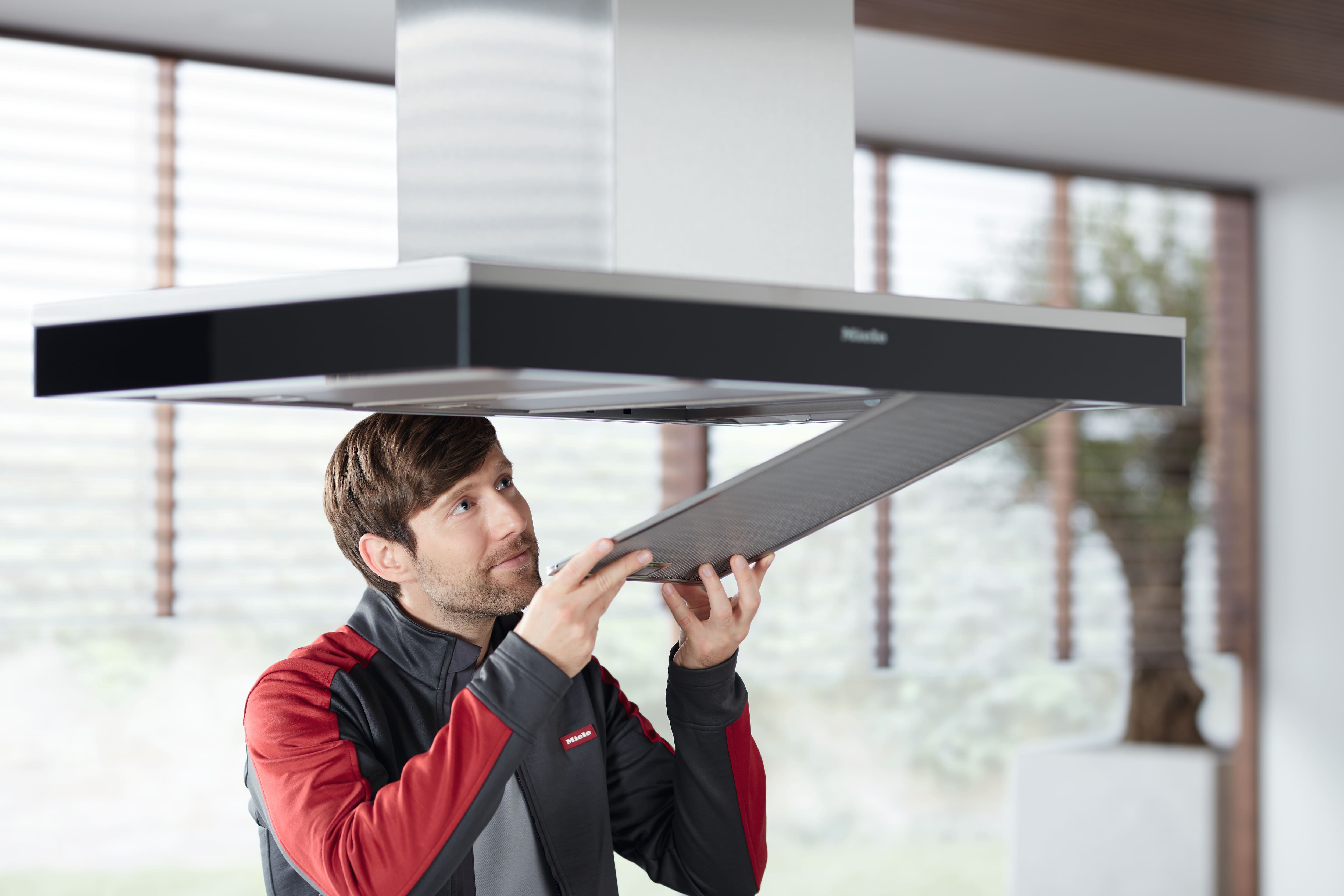 維修技師在明亮現代的廚房環境中，為 Miele 島式抽油煙機安裝網狀油脂過濾器。