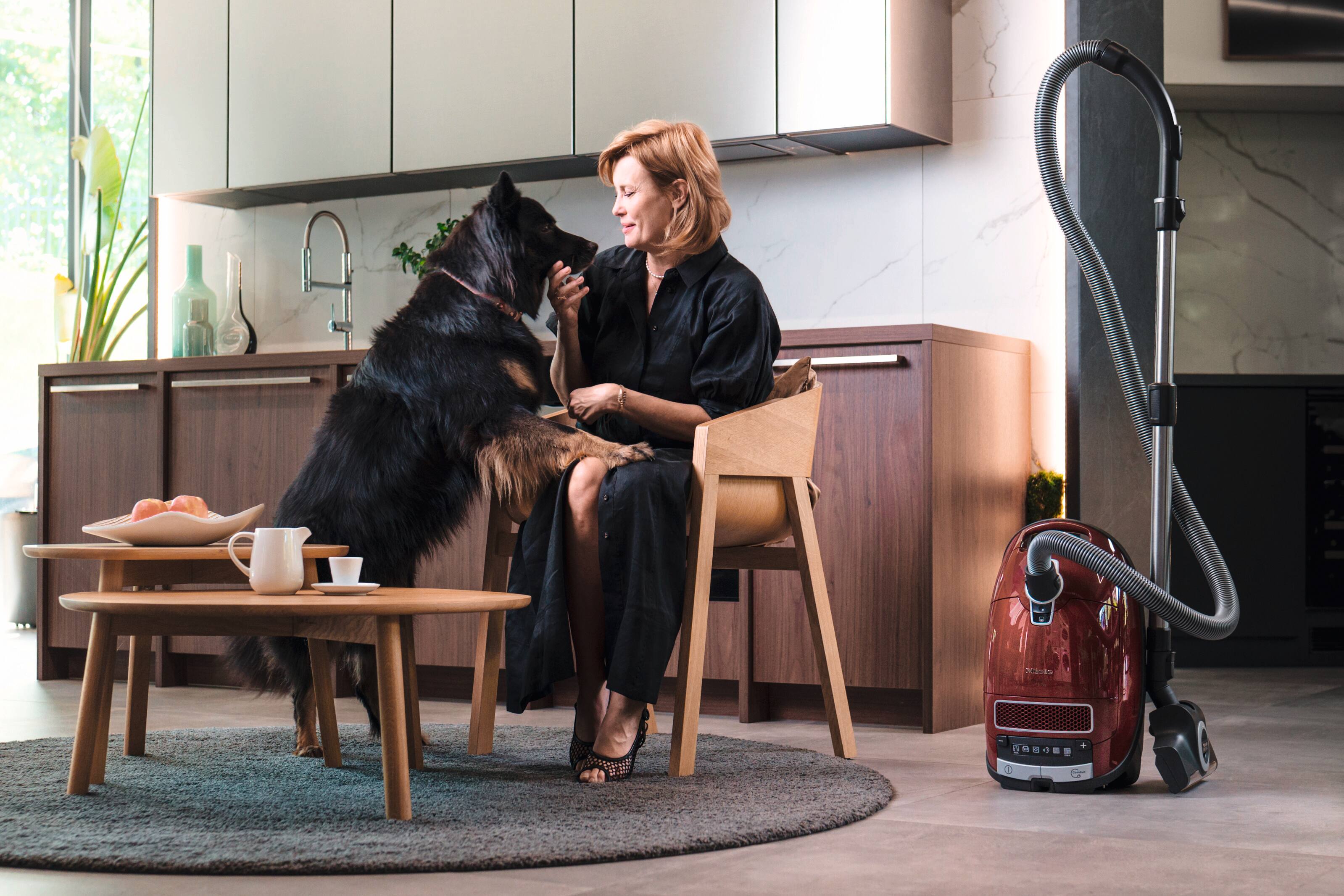 Cozy kitchen scene: a woman pets her large dog beside a red canister vacuum, coffee table, and wood cabinets. Cozy kitchen scene: a woman pets her large dog beside a red canister vacuum, coffee table, and wood cabinets.
