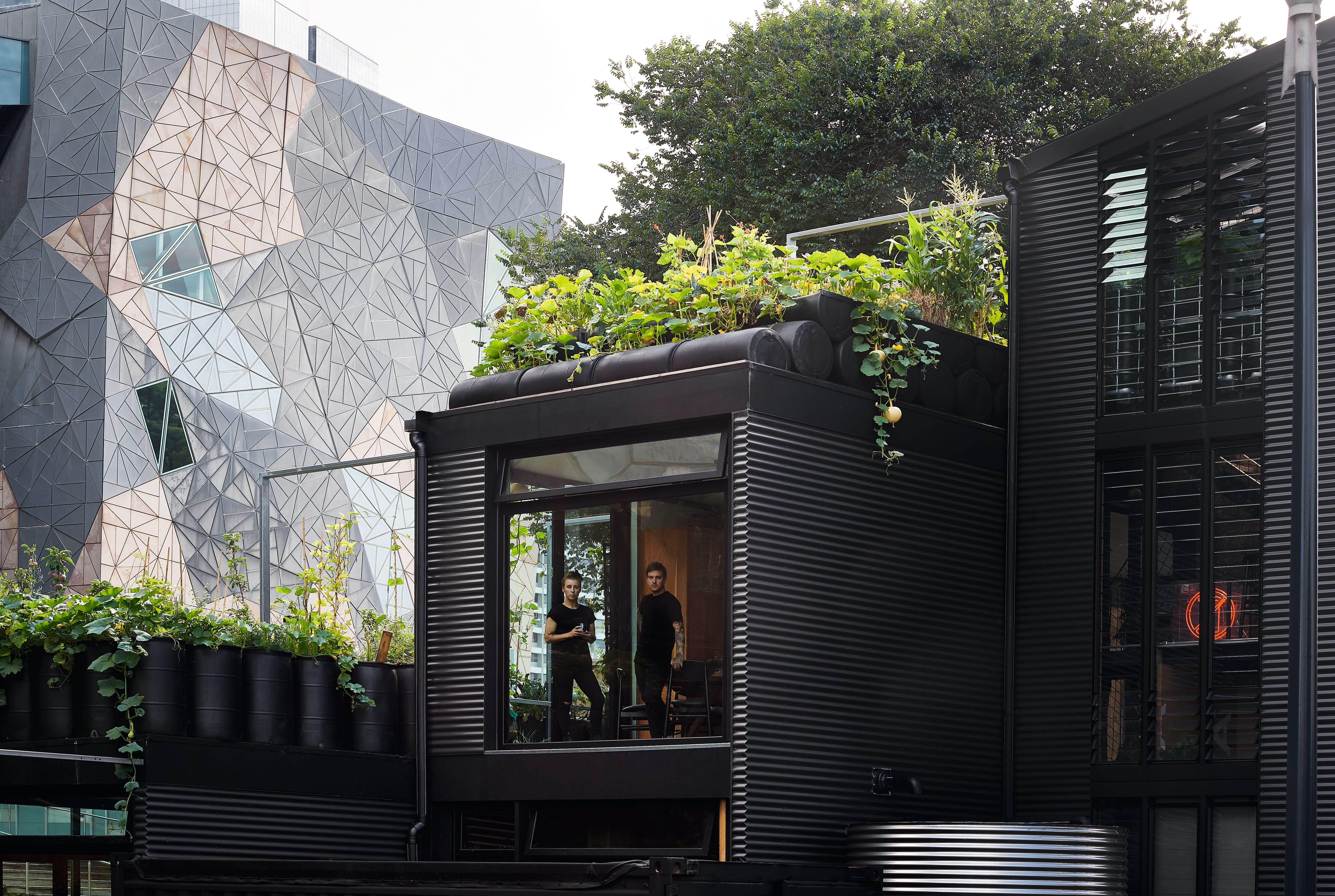 Edificio moderno nero ondulato con giardino sul tetto; due persone visibili all'interno; facciata geometrica urbana dietro. Edificio moderno nero ondulato con giardino sul tetto; due persone visibili all'interno; facciata geometrica urbana dietro.