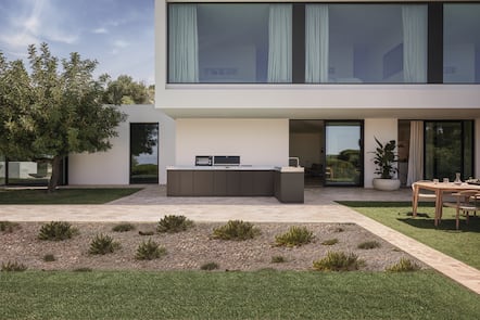 Modern home patio featuring a sleek outdoor kitchen with grill and sink, large windows, landscaped garden, and a wood dining set.