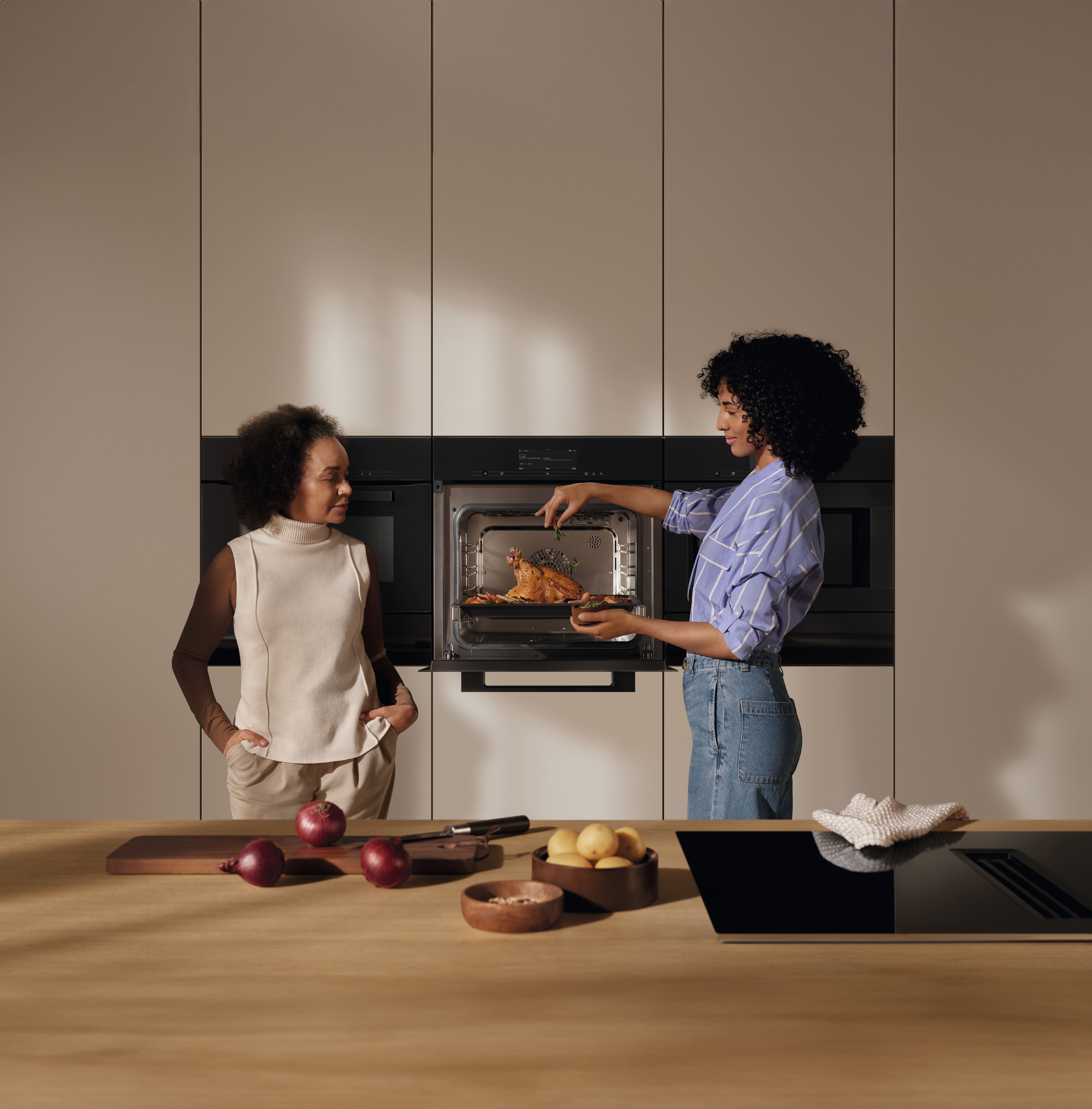Twee mensen in een moderne keuken halen een gebraden kip uit een inbouwoven, met uien en aardappelen op het aanrecht.