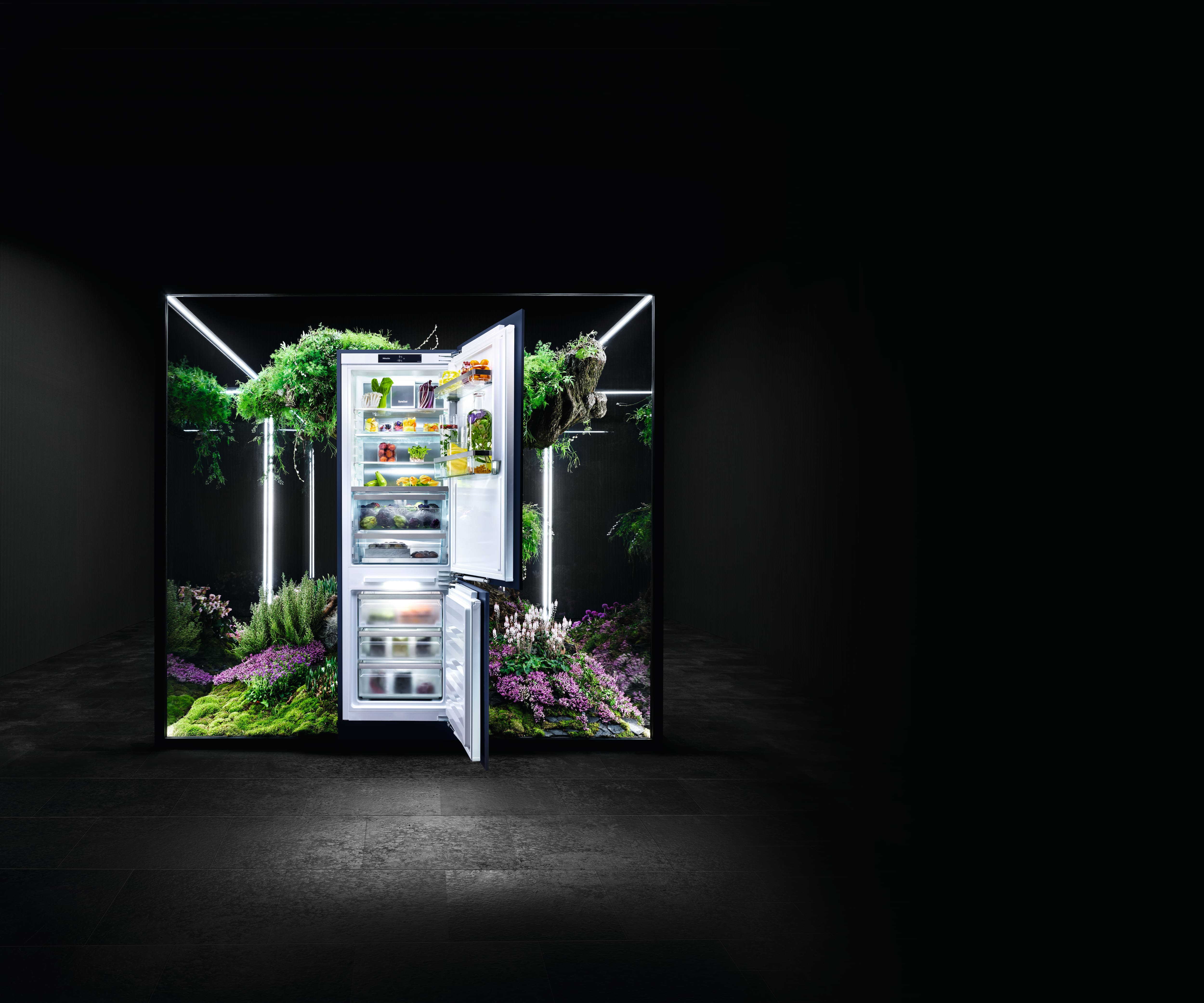 Open, illuminated Miele refrigerator in a glass cube surrounded by lush greenery and flowers, contrasting a dark room. Open, illuminated Miele refrigerator in a glass cube surrounded by lush greenery and flowers, contrasting a dark room.