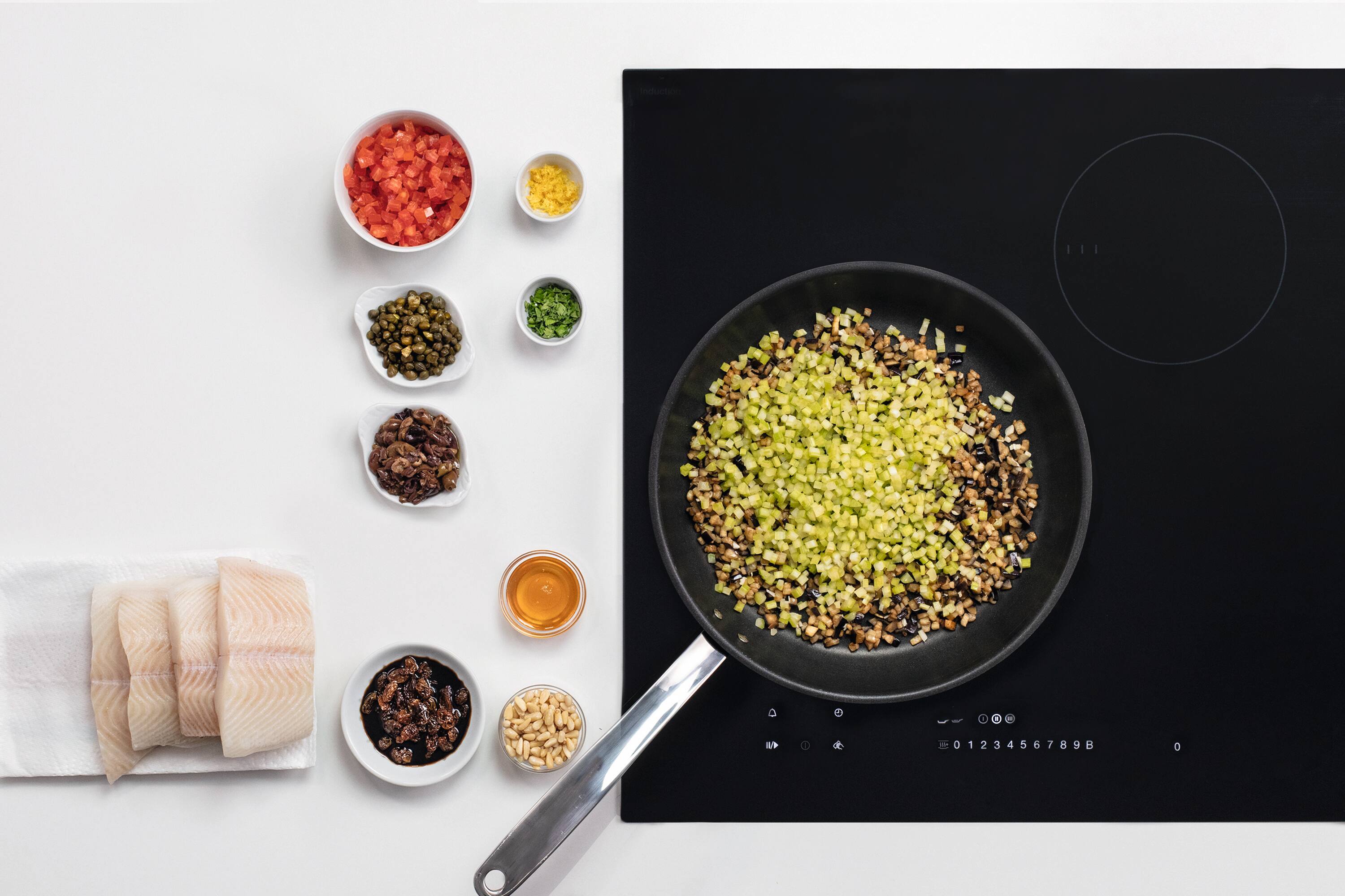 Overhead view of an induction cooktop: pan with diced veg; bowls of tomatoes, capers, nuts, and fish fillets. Overhead view of an induction cooktop: pan with diced veg; bowls of tomatoes, capers, nuts, and fish fillets.