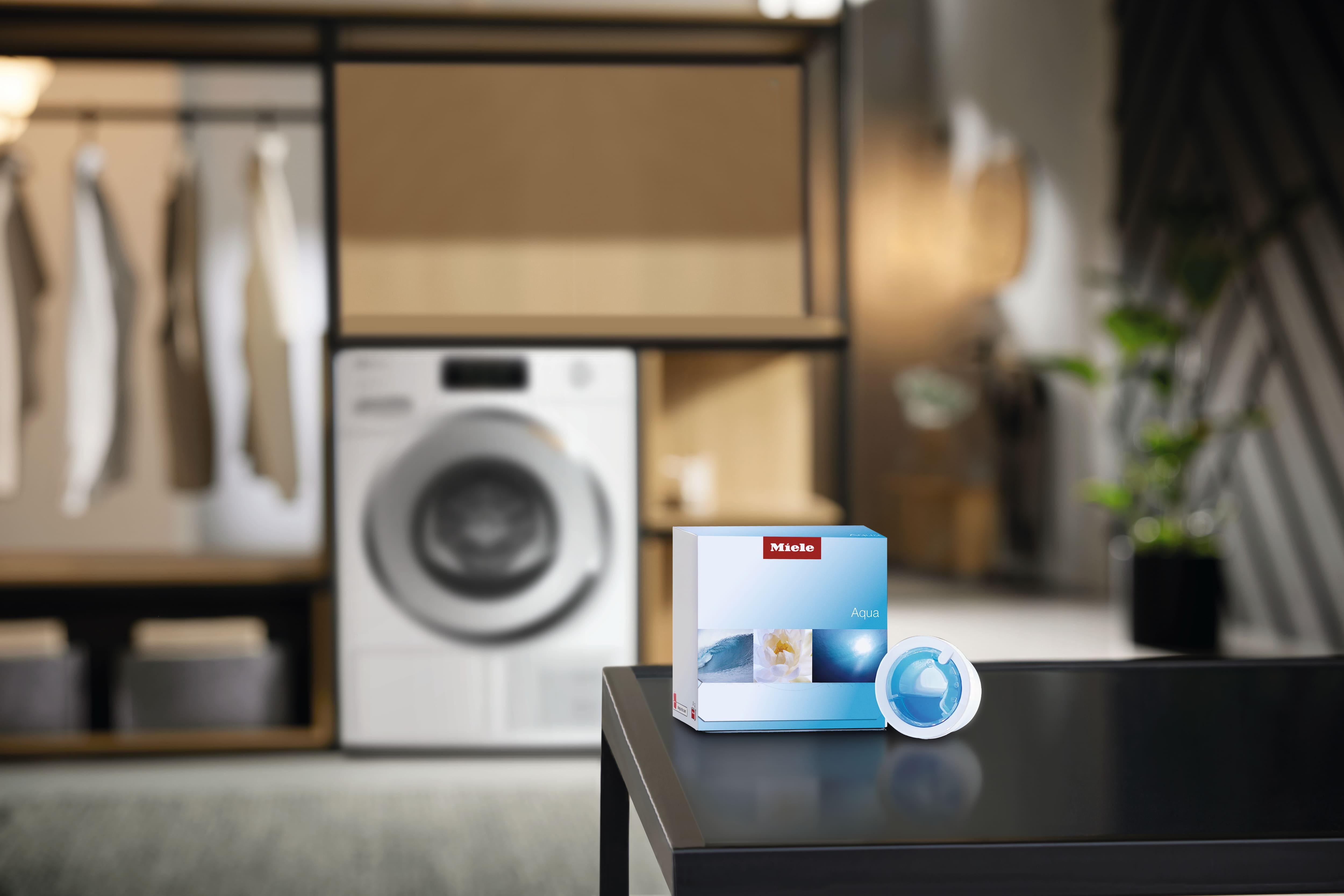 Box of Miele Aqua detergent pods and single pod on table in modern laundry room with washer, highlighting freshness. Box of Miele Aqua detergent pods and single pod on table in modern laundry room with washer, highlighting freshness.