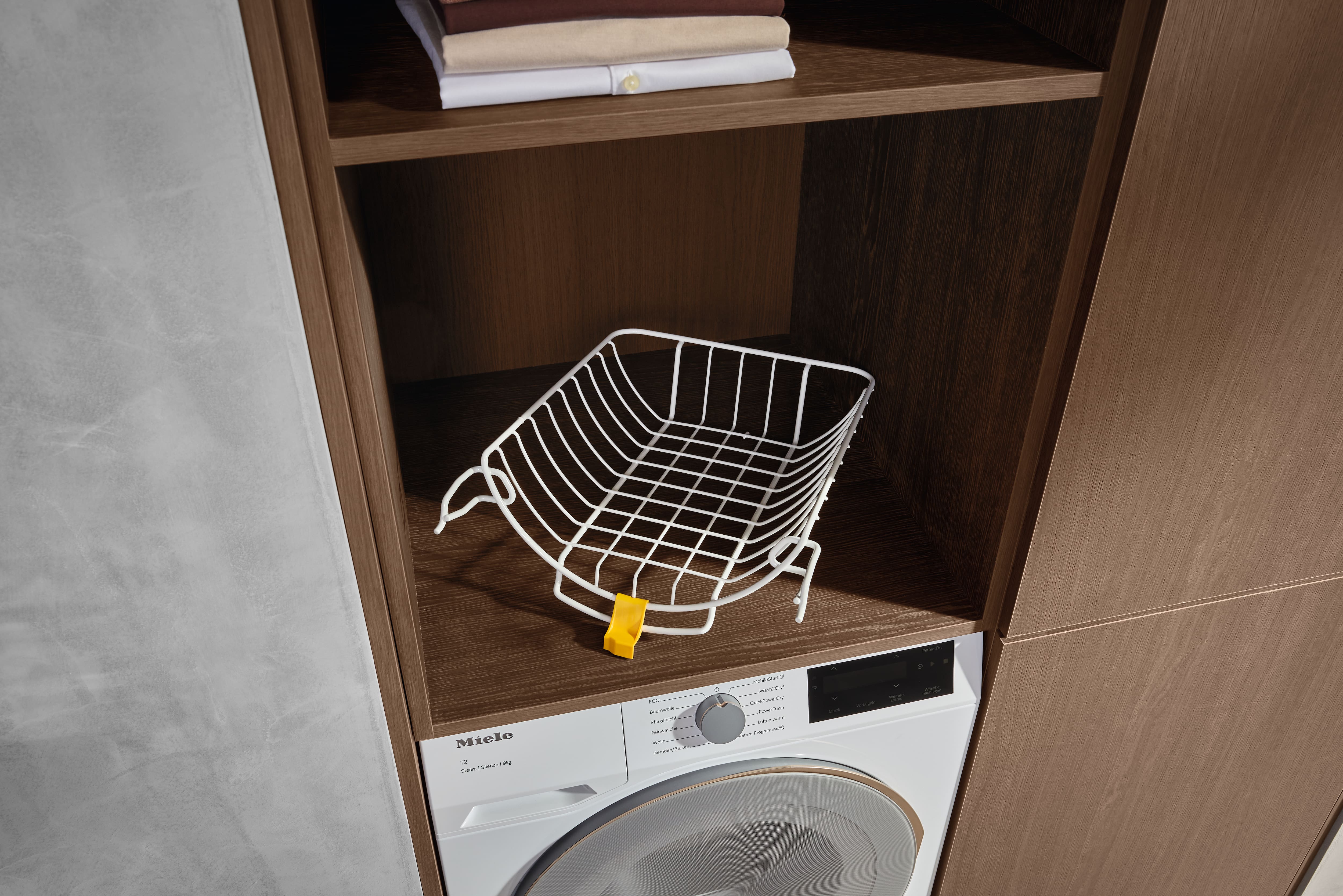 White wire laundry basket on a wooden shelf in a built-in cabinet above a Miele dryer, folded shirts above. White wire laundry basket on a wooden shelf in a built-in cabinet above a Miele dryer, folded shirts above.
