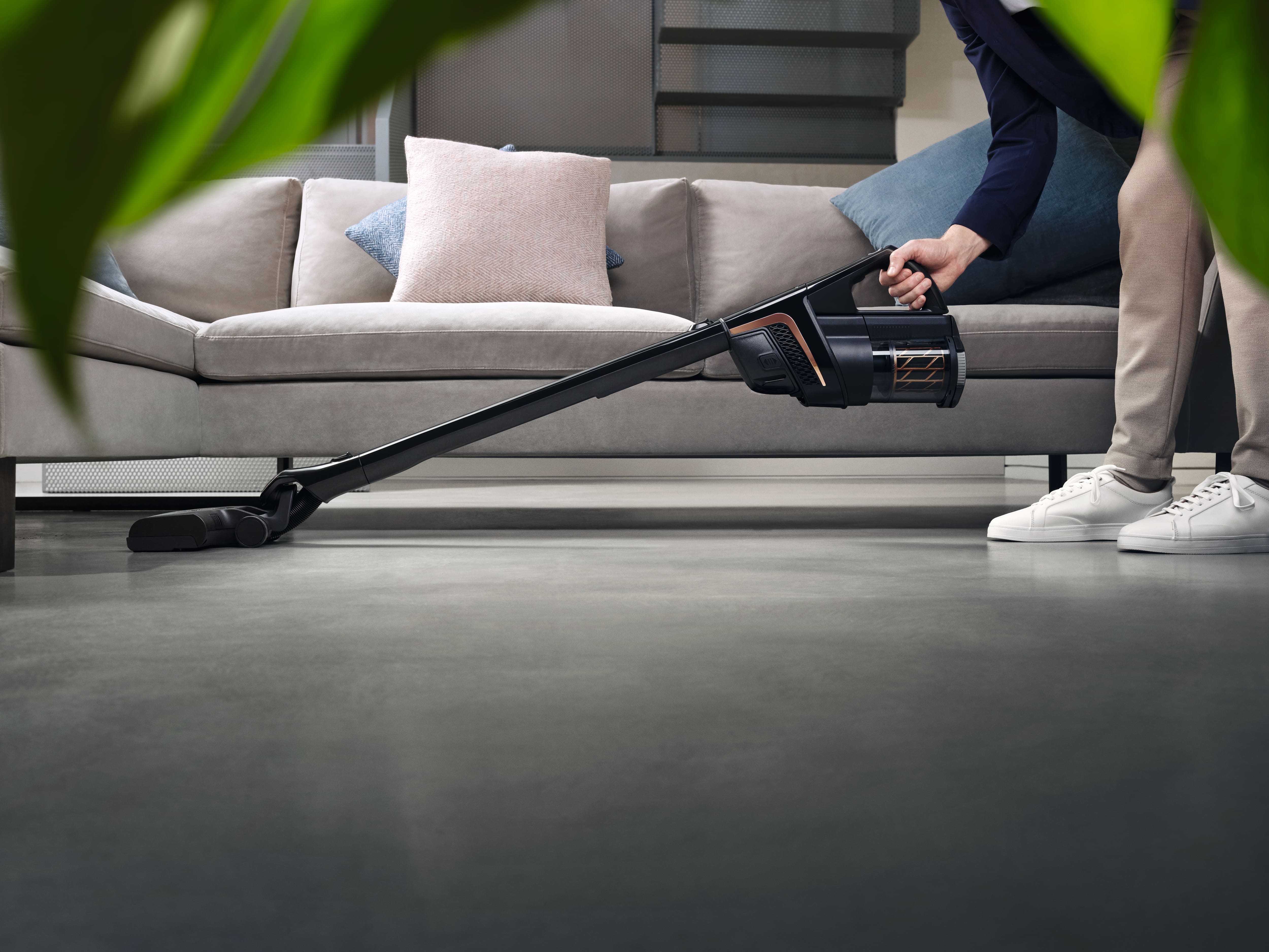Person using a cordless stick vacuum to clean under a sofa on a hard floor in a modern living room.