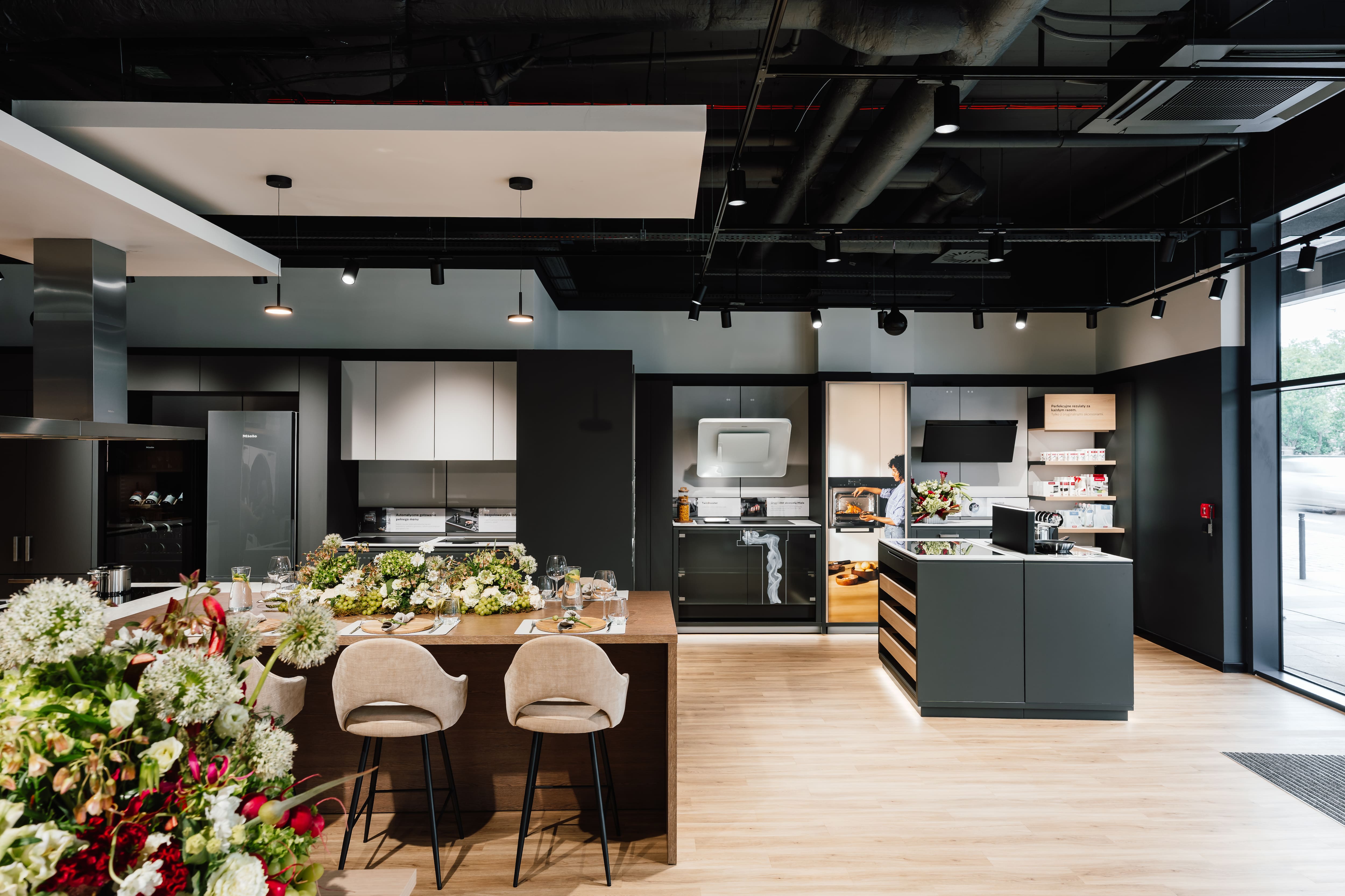 Modern showroom kitchen with island seating, sleek appliances, and floral table settings under track lighting. Modern showroom kitchen with island seating, sleek appliances, and floral table settings under track lighting.
