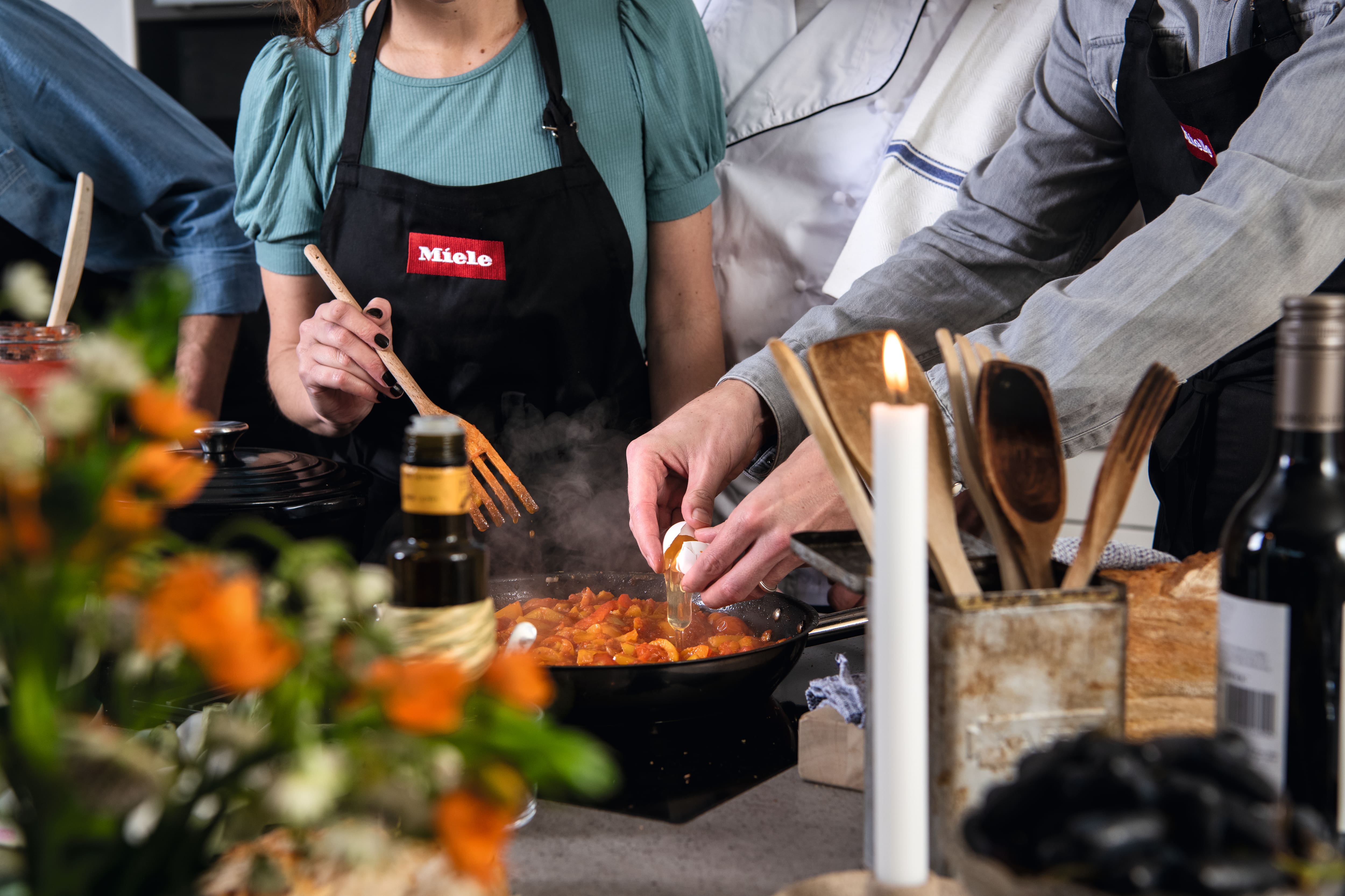 Group cooking scene: people in aprons crack an egg into a steaming pan, with utensils, candle, wine, and a Miele logo.
