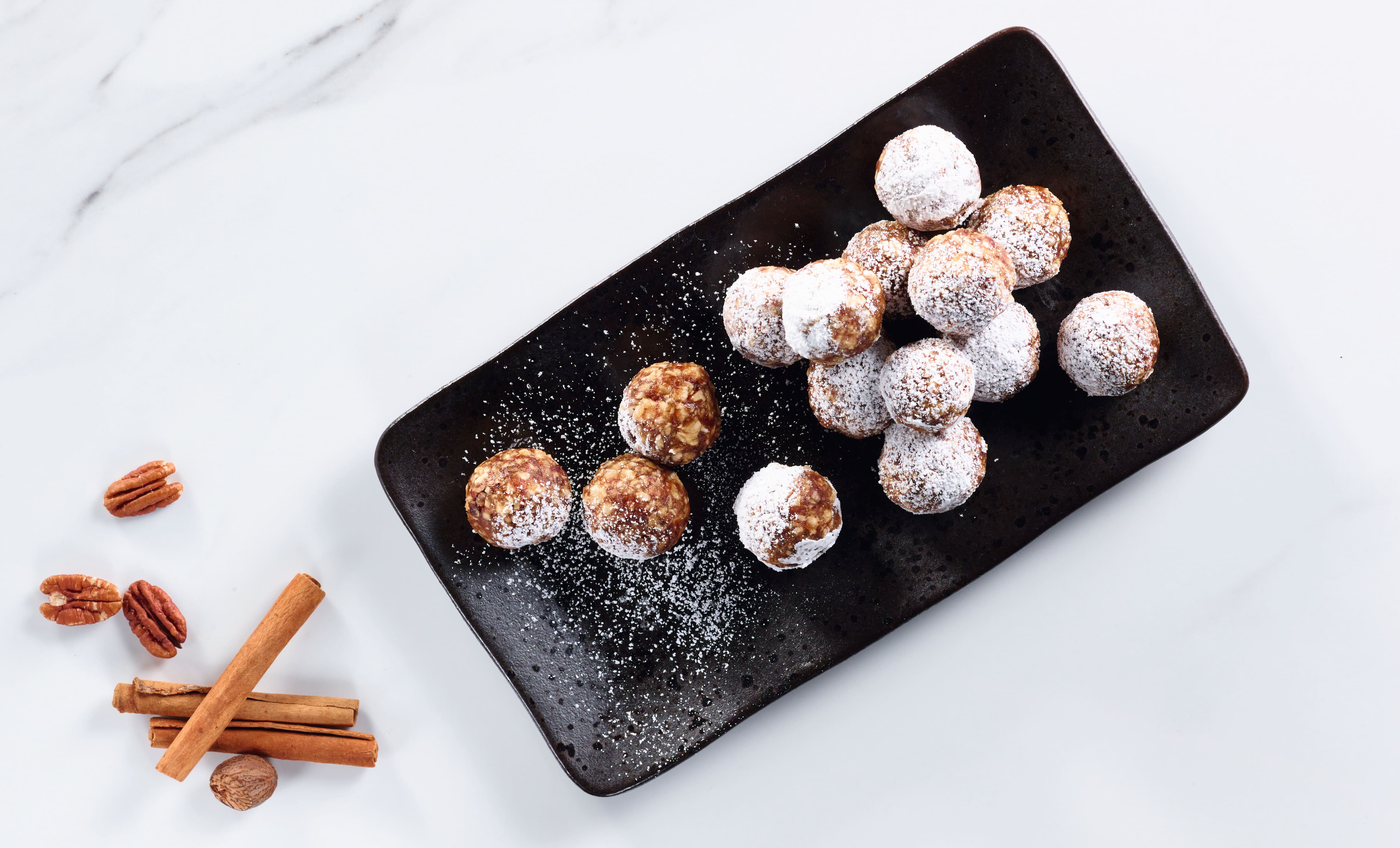 Black rectangular plate with small round dessert balls dusted in powdered sugar, on a white marble surface; cinnamon sticks, pecans, and a whole nutmeg are placed beside the plate. Black rectangular plate with small round dessert balls dusted in powdered sugar, on a white marble surface; cinnamon sticks, pecans, and a whole nutmeg are placed beside the plate.