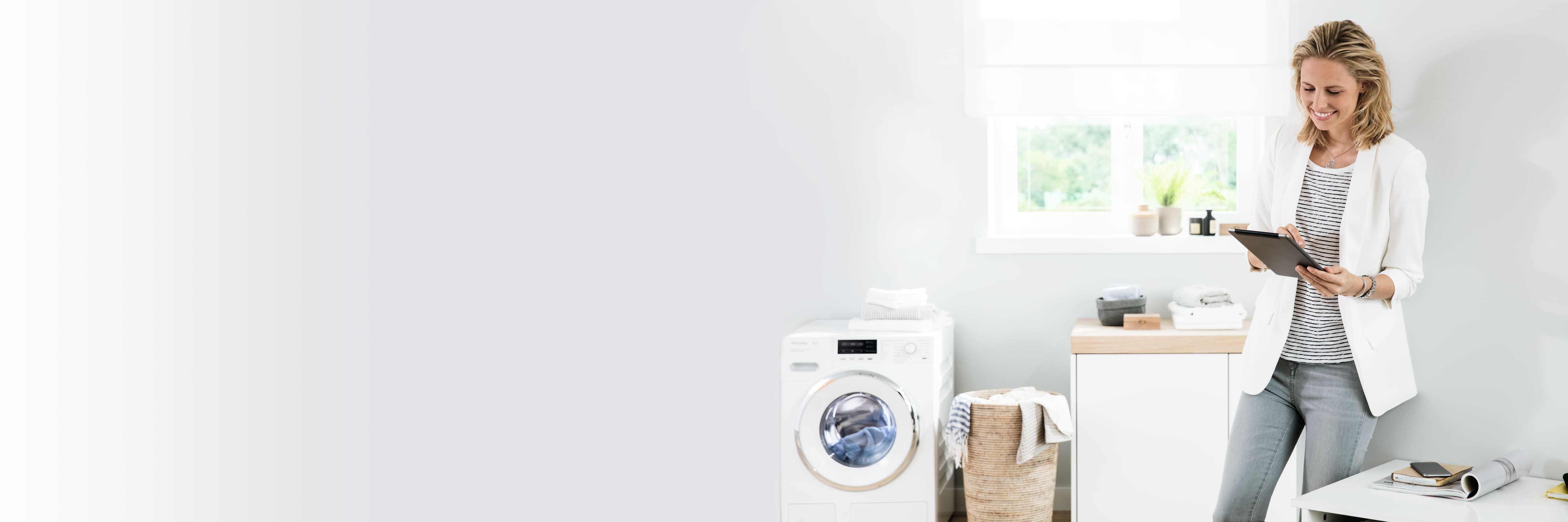 Bright laundry room with washer, wicker basket, and folded towels; a person in a white blazer uses a tablet by the window. Bright laundry room with washer, wicker basket, and folded towels; a person in a white blazer uses a tablet by the window.