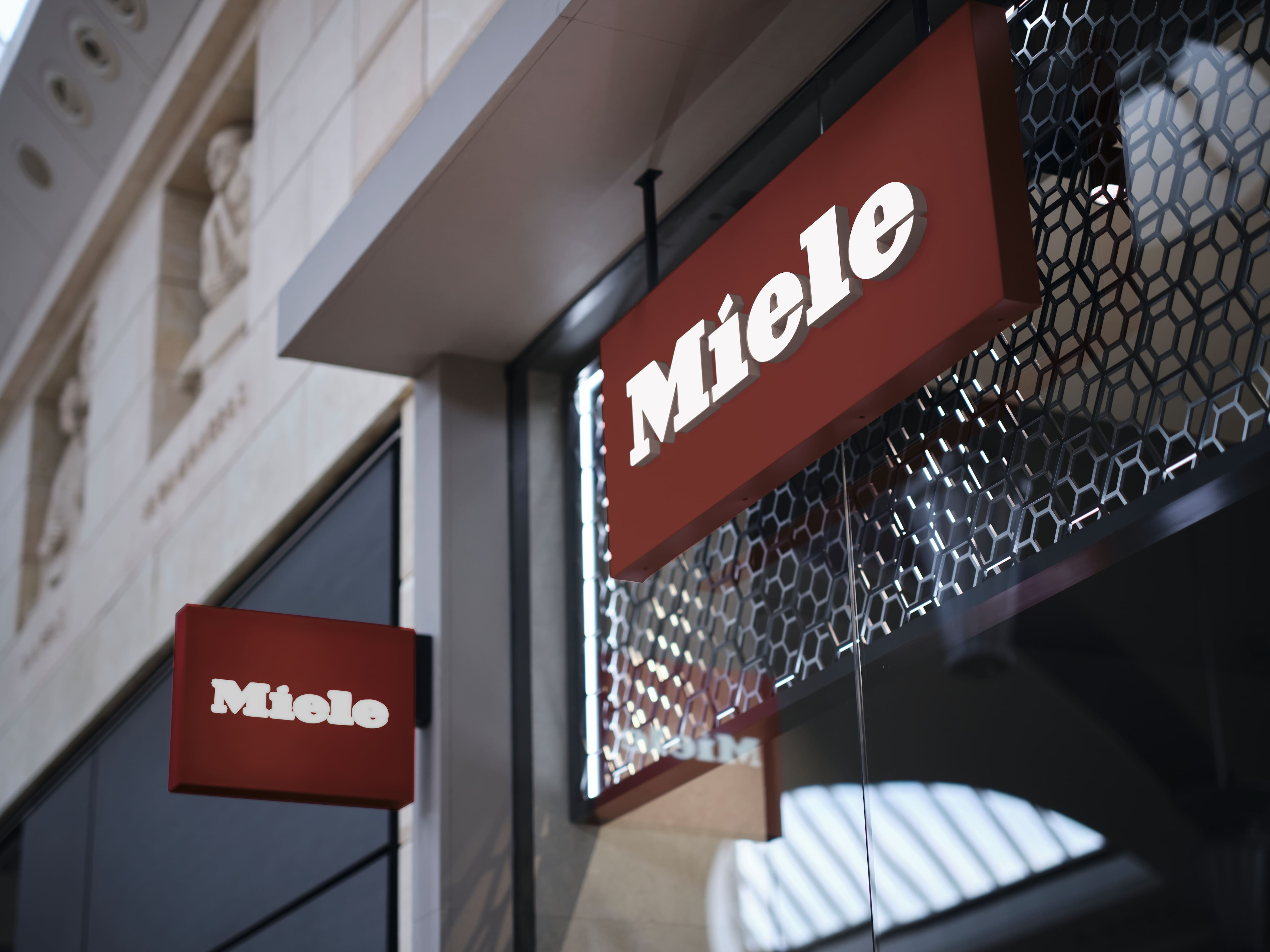 Red storefront signs with white 'Miele' lettering on a modern facade, showcasing brand presence at a retail location.