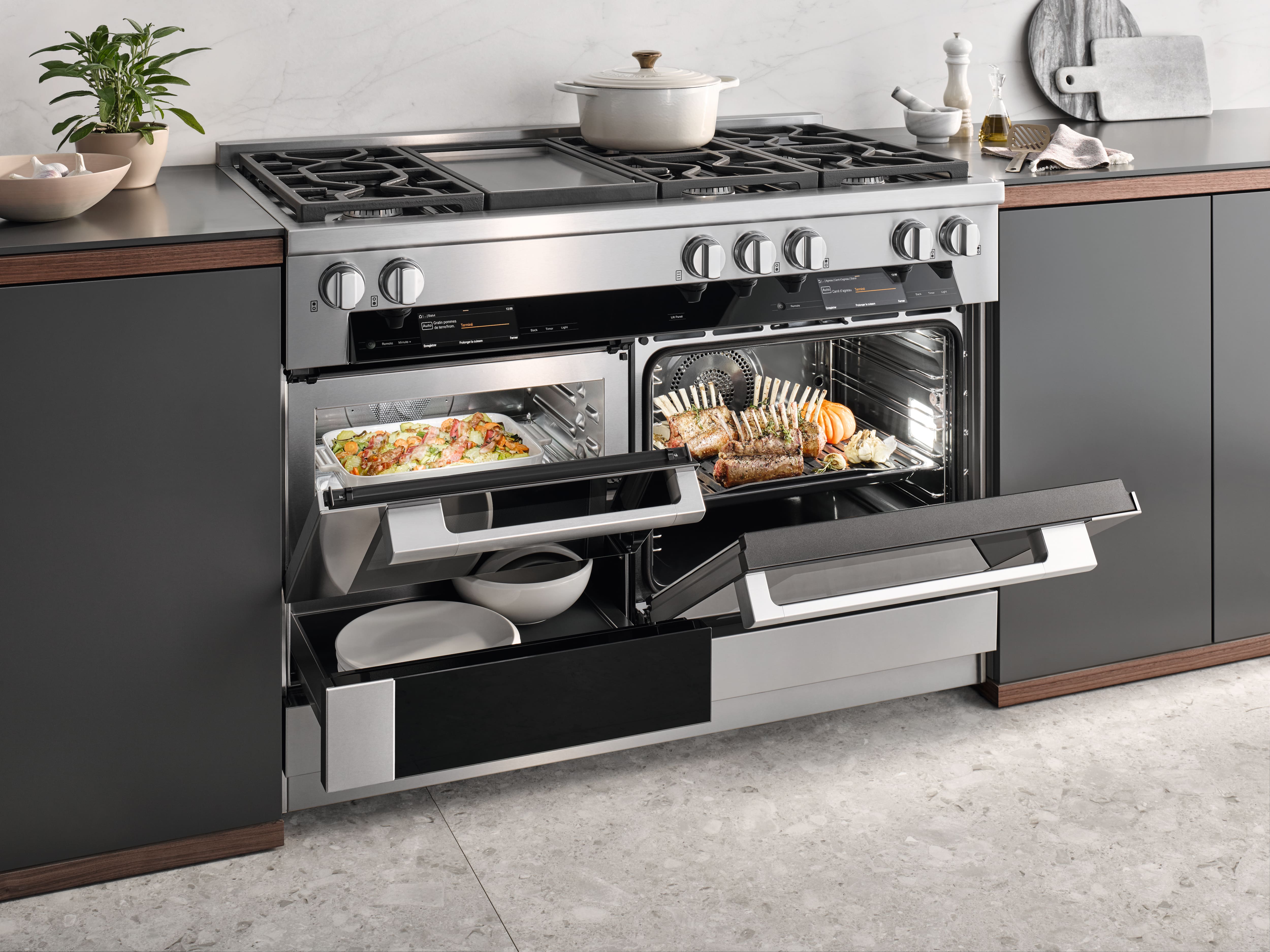 Modern stainless-steel range with gas cooktop and open ovens roasting lamb and a casserole in a sleek kitchen.