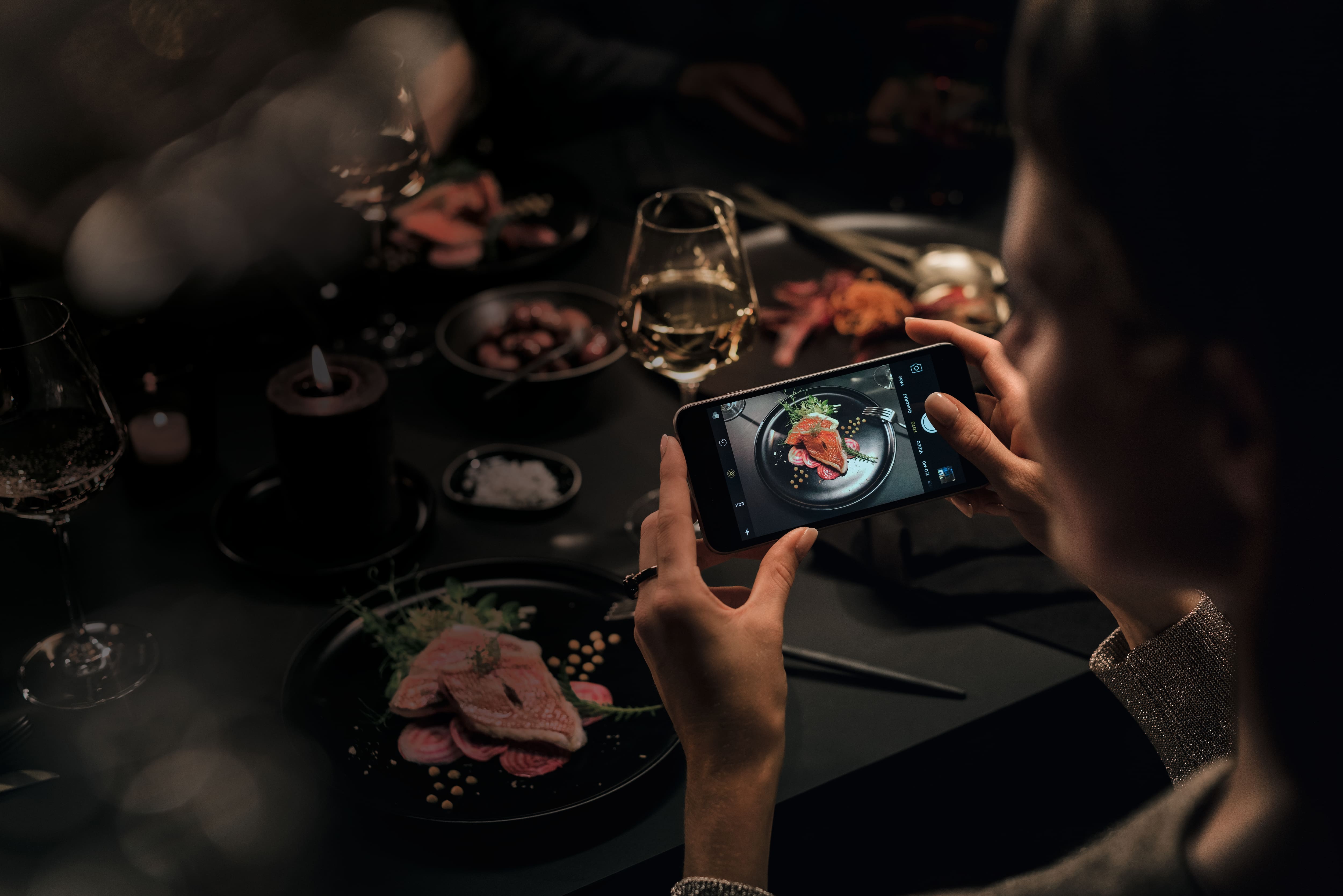 Osoba fotografująca smartfonem wykwintne danie na talerzu przy słabo oświetlonym stoliku w restauracji, przy świecach i kieliszku wina.