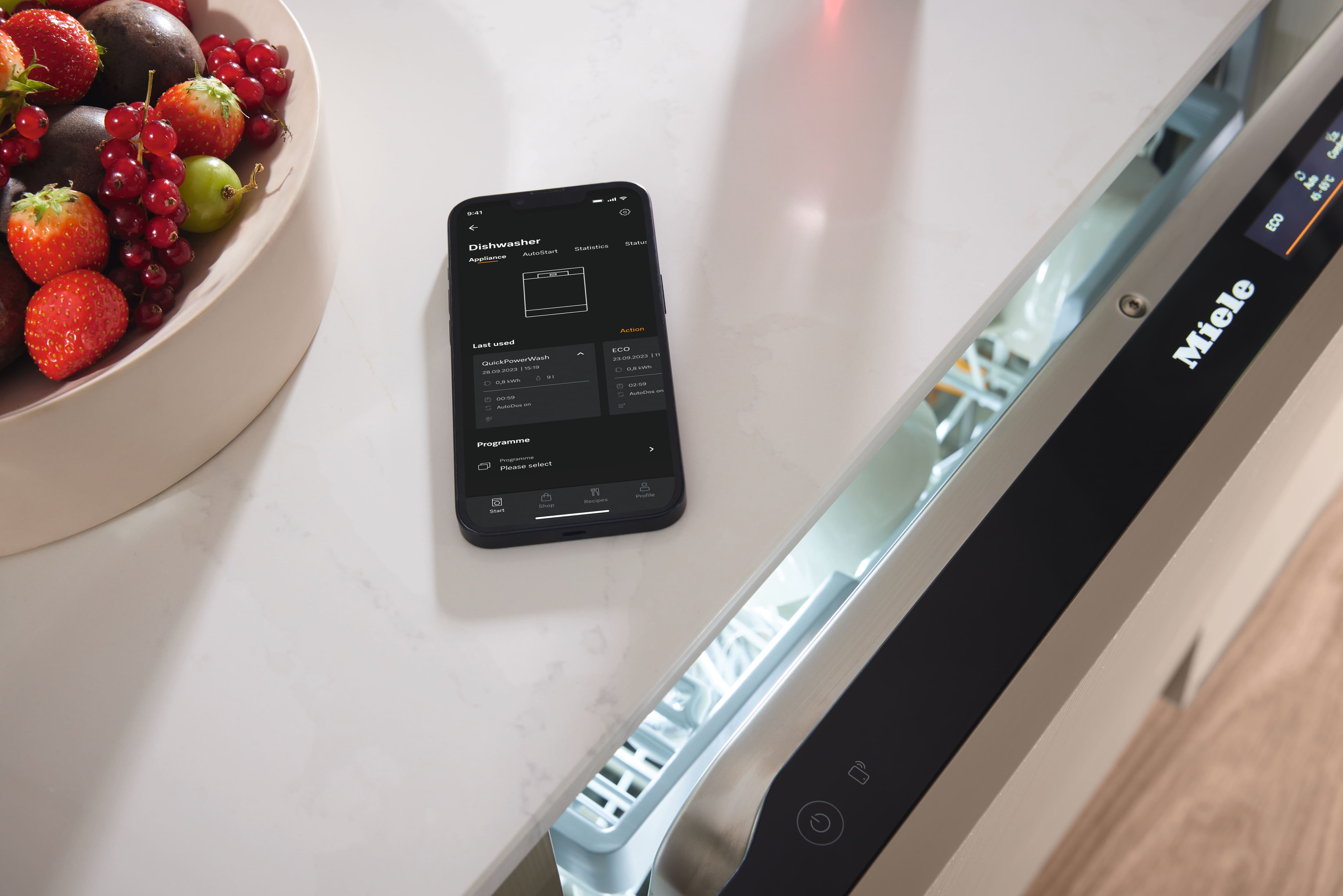 Smartphone displaying dishwasher control app on kitchen counter beside fruit bowl and open Miele dishwasher. Smartphone displaying dishwasher control app on kitchen counter beside fruit bowl and open Miele dishwasher.