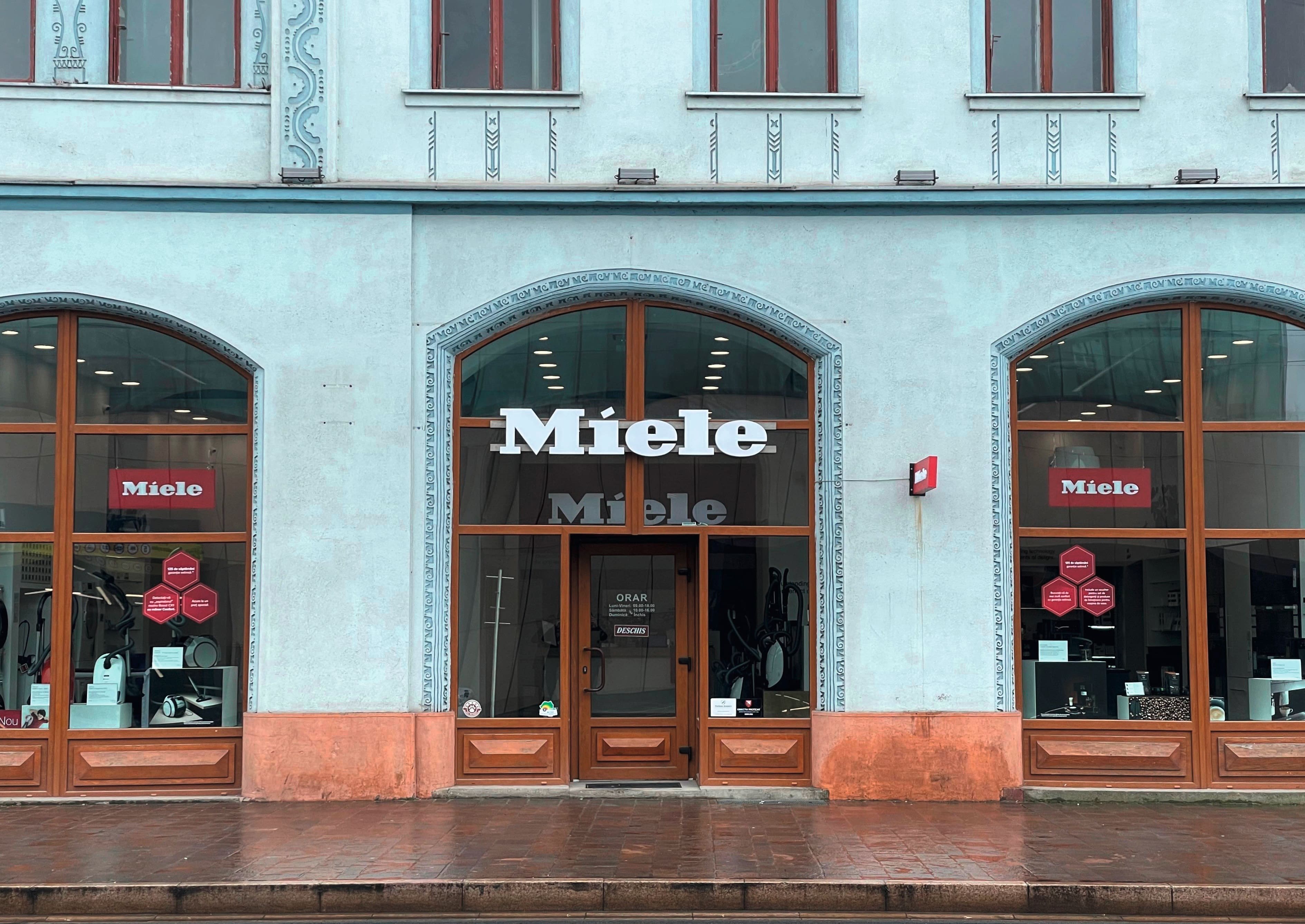 Vedere stradală a unui magazin de electrocasnice Miele cu ferestre mari arcuite, rame din lemn și trotuar umed după ploaie. Vedere stradală a unui magazin de electrocasnice Miele cu ferestre mari arcuite, rame din lemn și trotuar umed după ploaie.
