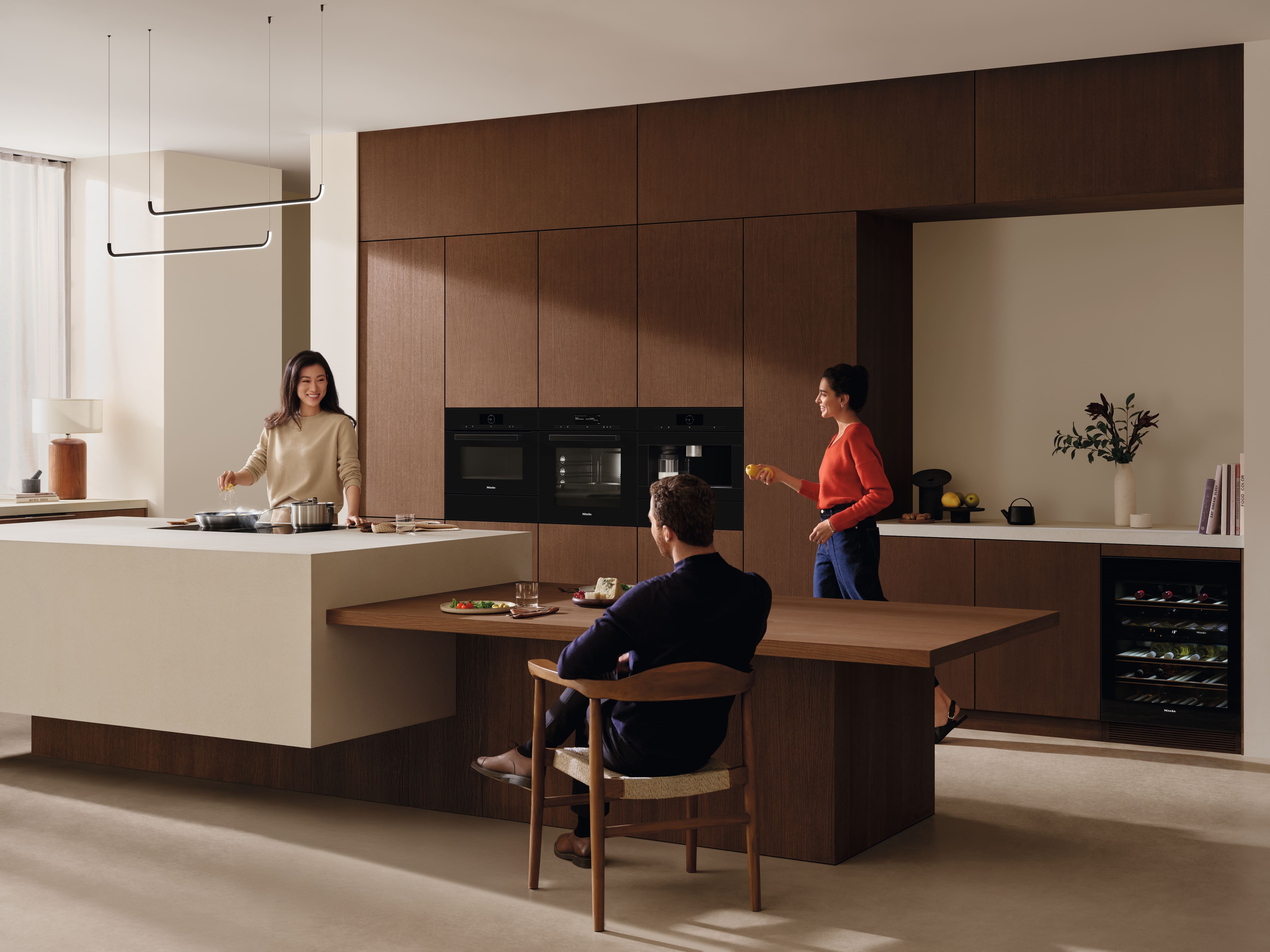 Cocina moderna con cálidos armarios de madera; tres personas cocinando y charlando alrededor de la isla y electrodomésticos integrados. Cocina moderna con cálidos armarios de madera; tres personas cocinando y charlando alrededor de la isla y electrodomésticos integrados.